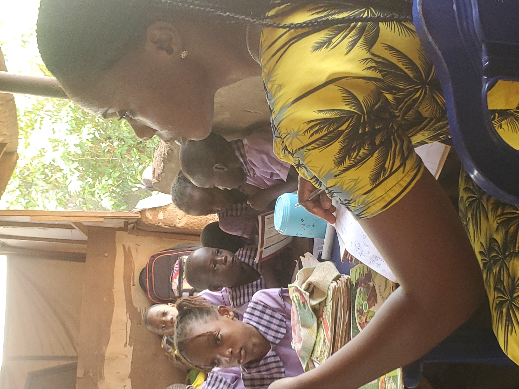 In Ukwu-Abwa, Nigeria, a group of children are gathered inside a structure with earthen walls and a corrugated metal roof. A woman wearing a yellow palm tree patterned shirt is leaning over them, holding a pen and paper, seemingly instructing them. The children are wearing striped shirts, and several of them are looking at books and writing. A backpack sits against the wall behind them. Light enters through a window or opening in the wall.