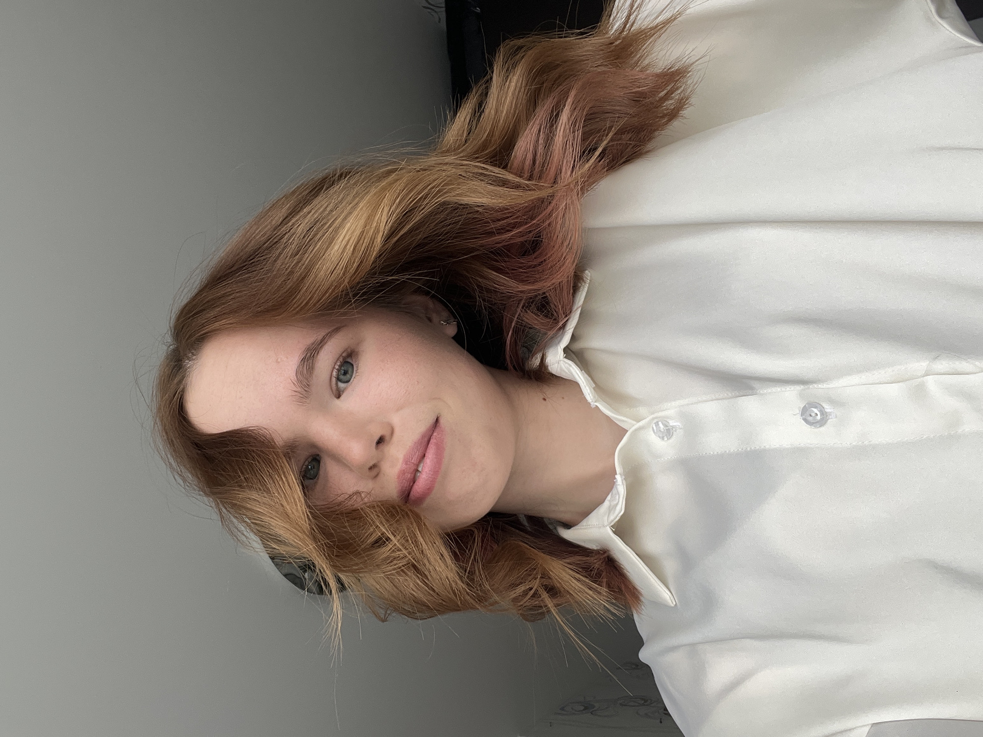 A young woman with light skin and strawberry blonde hair is pictured indoors against a gray wall. Her hair is styled with some strands falling across her face. She is wearing a white button-down shirt. No information suggests this image was taken in Chaikovsky, Russia.