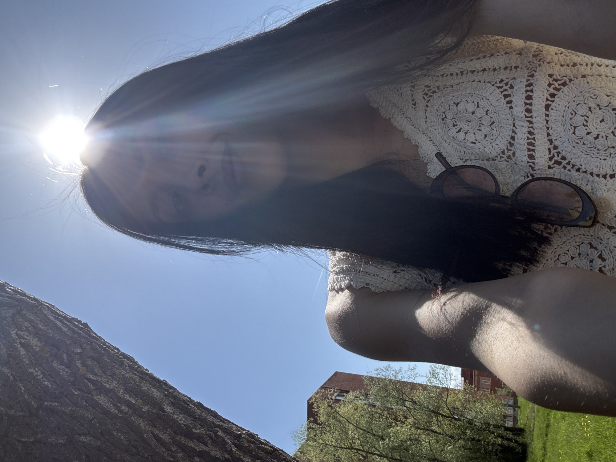 A young woman is seen from a low angle in an outdoor setting. She has long, dark hair and is wearing a crocheted top. A pair of sunglasses is clipped to her top. The bright sun is behind her head, causing lens flare. A tree trunk is visible on the left side of the frame. In the background, there is a building with a red roof, some trees, and green grass. The location is presumed to be in Votkinsk, Russia.