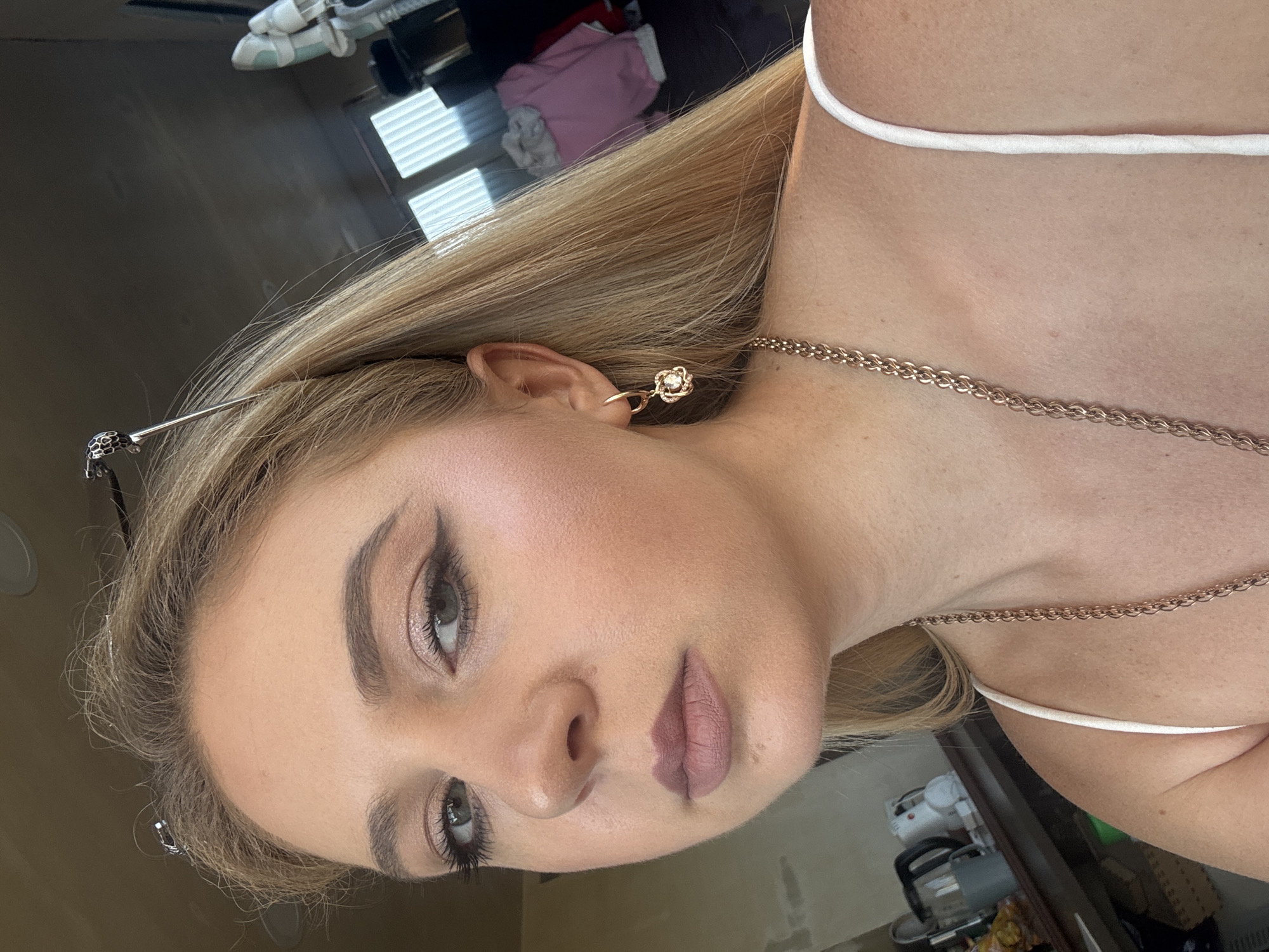 The image shows a close-up, vertically oriented portrait of a young woman. She has blonde hair and is wearing makeup, including eyeliner and lipstick. She has a delicate gold earring in her left ear and a rose gold chain necklace. The lighting suggests it might be daytime, possibly indoors given the blinds visible in the background. Her skin appears smooth and lightly tanned. The woman's expression is neutral, and the focus is on her face and upper body. The background is blurred, but hints of furniture and other objects are discernible, suggesting an indoor environment.