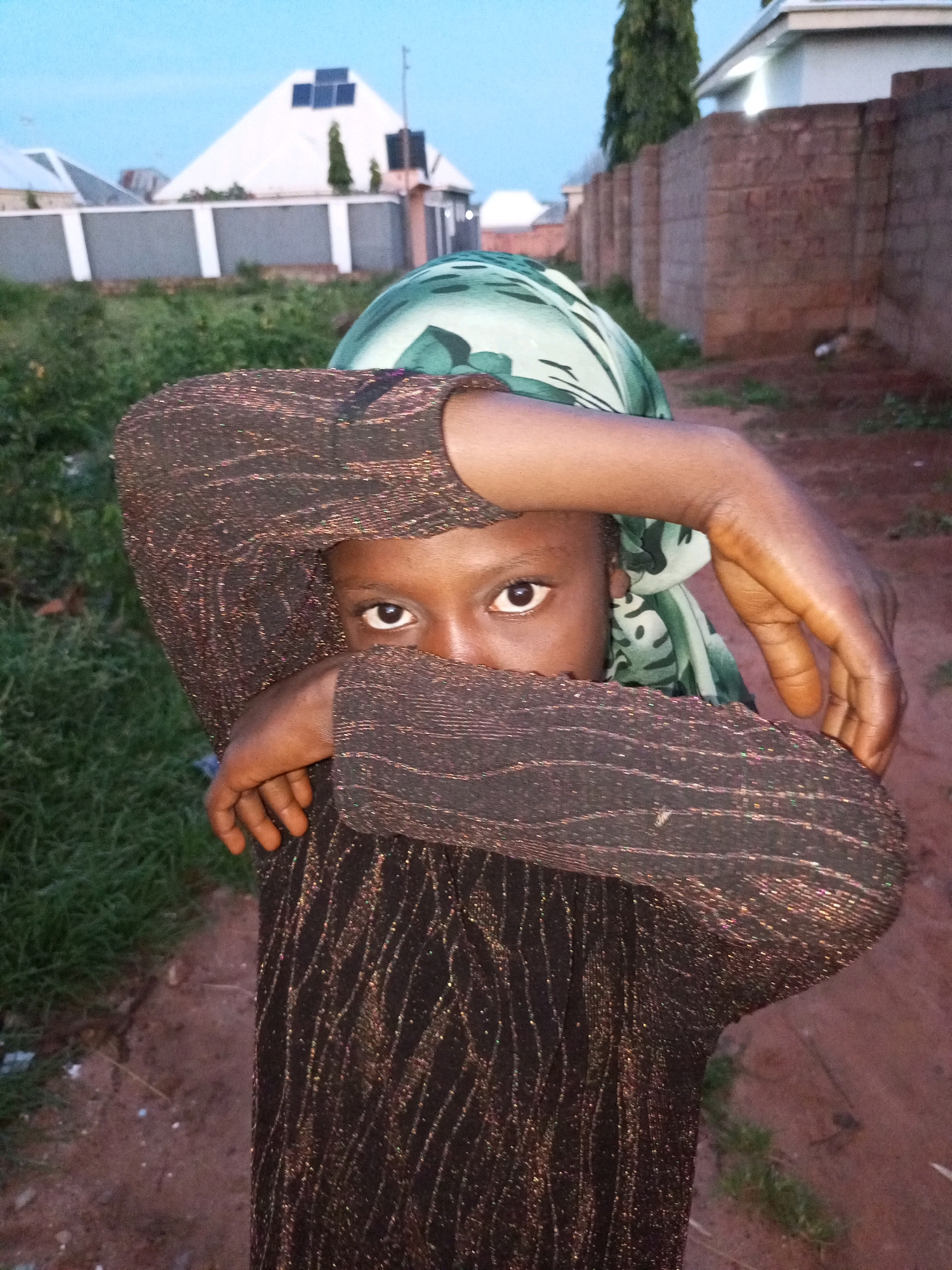The image shows a close-up of a young Black individual wearing a dark, sparkling long-sleeved garment and a green and white patterned headscarf. Their face is partially obscured by their arms, with only one eye visible. The background consists of a dirt path, green grass, and several buildings, including one with a triangular roof and solar panels. The lighting suggests it is dusk. The specific location is in the city of Tempure, Nigeria.