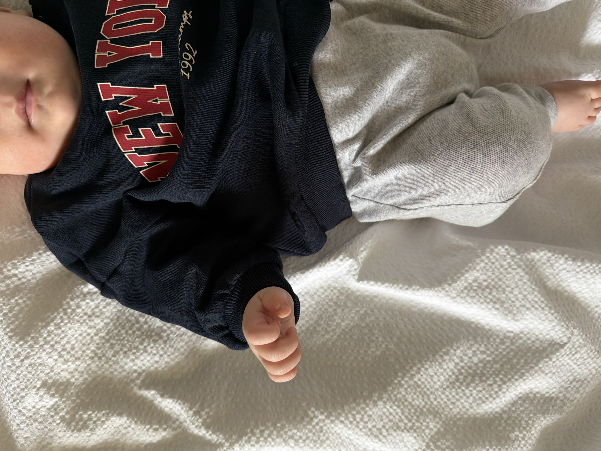An overhead shot captures a baby, likely an infant, peacefully resting on a white, textured blanket or sheet. The baby is dressed in a navy blue long-sleeved top and light grey pants. Visible are the lower portion of the baby's face, including the mouth and chin, a small, partially clenched hand on the left side, and a bare foot on the right.
The navy blue top features prominent red text with a white outline, spelling "NEW YORK". Below this, smaller, lighter script reads "Est. 1997". The clothing appears soft and comfortable, with visible texture on the fabrics.
The scene is illuminated by natural light, suggesting daytime, casting soft shadows across the white blanket. The blanket itself has a distinct, waffle-like knit pattern. The overall mood of the image is calm and serene. While the location context is Khimki, Russia, there are no specific visual cues within the image itself to confirm this.