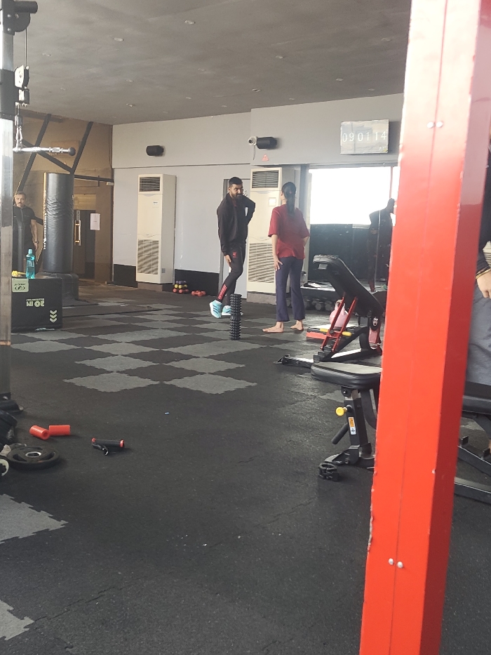 A gym setting shows a man in black attire standing on a foam roller, conversing with a woman wearing a red shirt and blue pants. Another man stands near a punching bag. Gym equipment is scattered around the black and gray checkered floor. A digital clock displays "09 01 14" on the wall. A red structural beam is partially visible in the foreground.