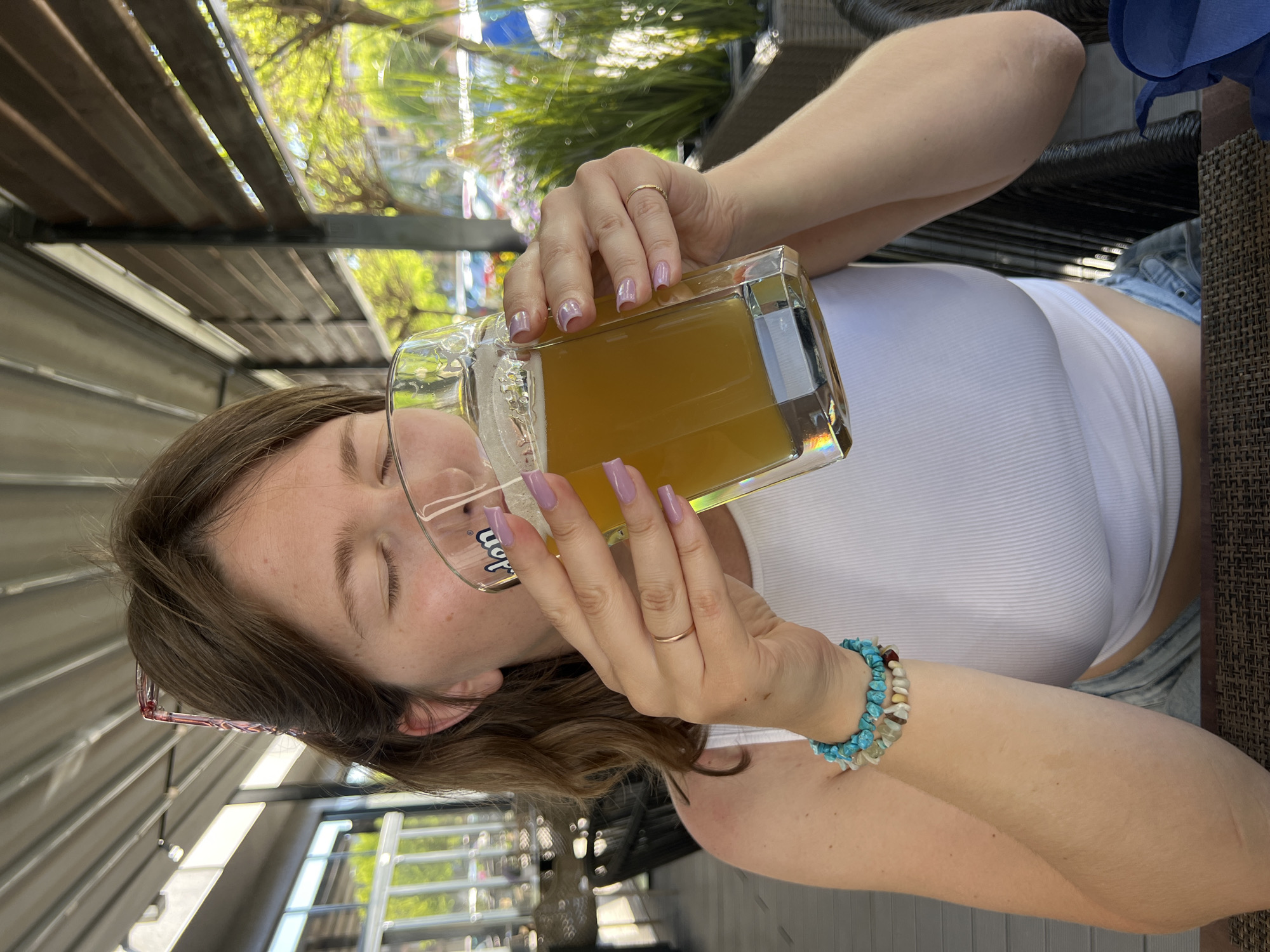 A young woman is drinking from a large, square glass mug filled with a golden-colored liquid, likely beer. She wears a white tank top, denim shorts, and turquoise beaded bracelets on her right wrist. Her nails are painted with a light purple color, and she has a gold ring on her finger. She is sitting outdoors, possibly at a cafe or restaurant. The setting appears to be Novi Sad, Serbia, suggested by architectural details in the background, though this is not confirmed.