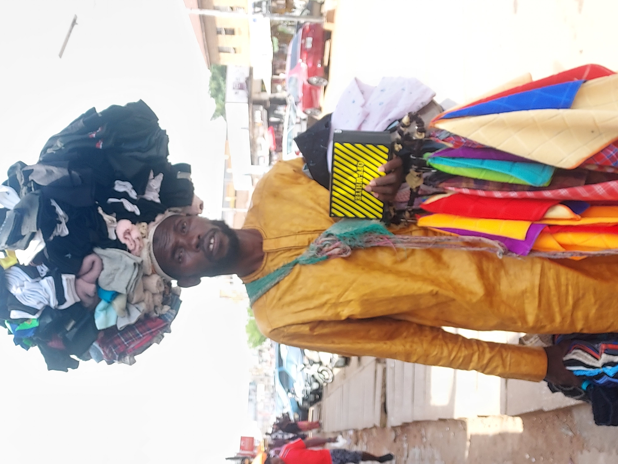 A man stands on a street in Jos, Nigeria, balancing a large bundle of clothing on his head and carrying more clothes and fabrics in his arms. He is wearing a mustard-yellow, long-sleeved shirt and is looking towards the viewer. The clothing on his head is a mixed assortment, while he holds colorful fabrics and what appears to be a black and yellow striped wallet or case in his right hand. A blue net is around his neck. The background shows a street scene with buildings, parked cars, a motorcycle, and a few people walking. The area appears to be a commercial or busy part of the city.