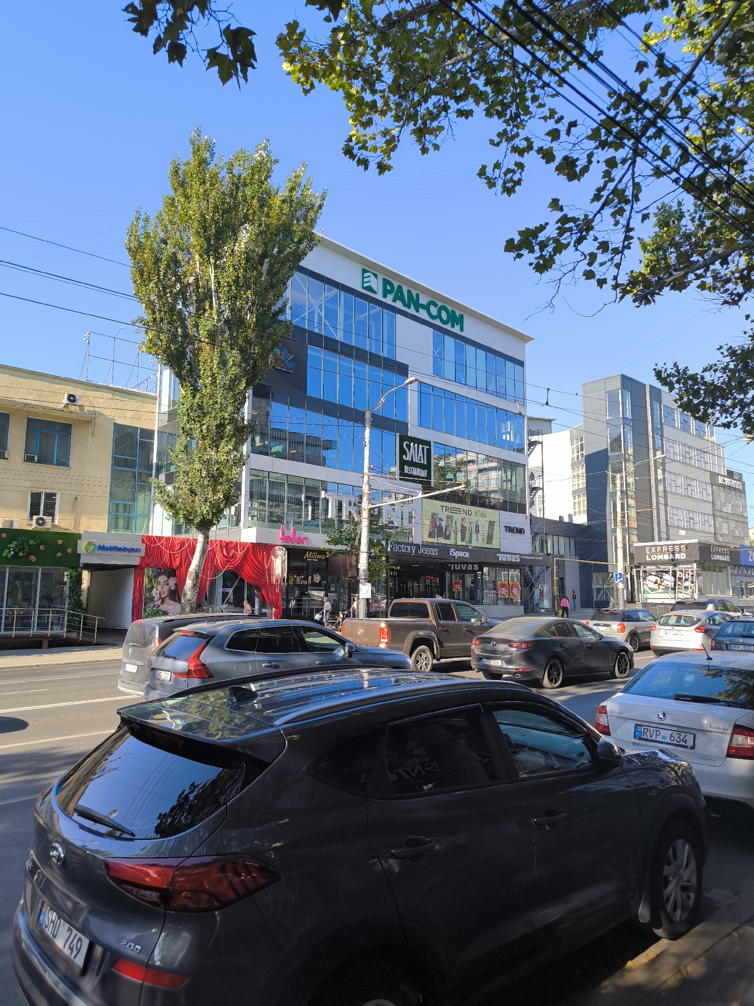 The image captures a street scene in Chișinău, Moldova, on a clear day. In the foreground, the rear of a dark gray Hyundai SUV is visible, with its license plate reading "SHD 749". To its right, a white Skoda Rapid with the license plate "RVP 634" is parked. Behind these vehicles, a dark gray Mazda sedan, a silver Volvo SUV with the license plate "ESBL 727", and a pickup truck are observed.
The background is dominated by a modern, multi-story building with a blue glass facade. A large green sign with white lettering that reads "PAN-COM" is prominent on the upper portion of this building. Below this, various shop signs are visible, including "SALAT RESTAURANT", "TREND Kids", "Factory Jeans", and "iSpace". Further to the right, another multi-story building with a gray facade is present, featuring commercial establishments such as "EXPRESS LOMBARD".
The street is lined with trees, one tall tree with green foliage directly obscures part of the "PAN-COM" building. Overhead power lines crisscross the sky. The overall environment suggests an urban commercial area with moderate traffic.