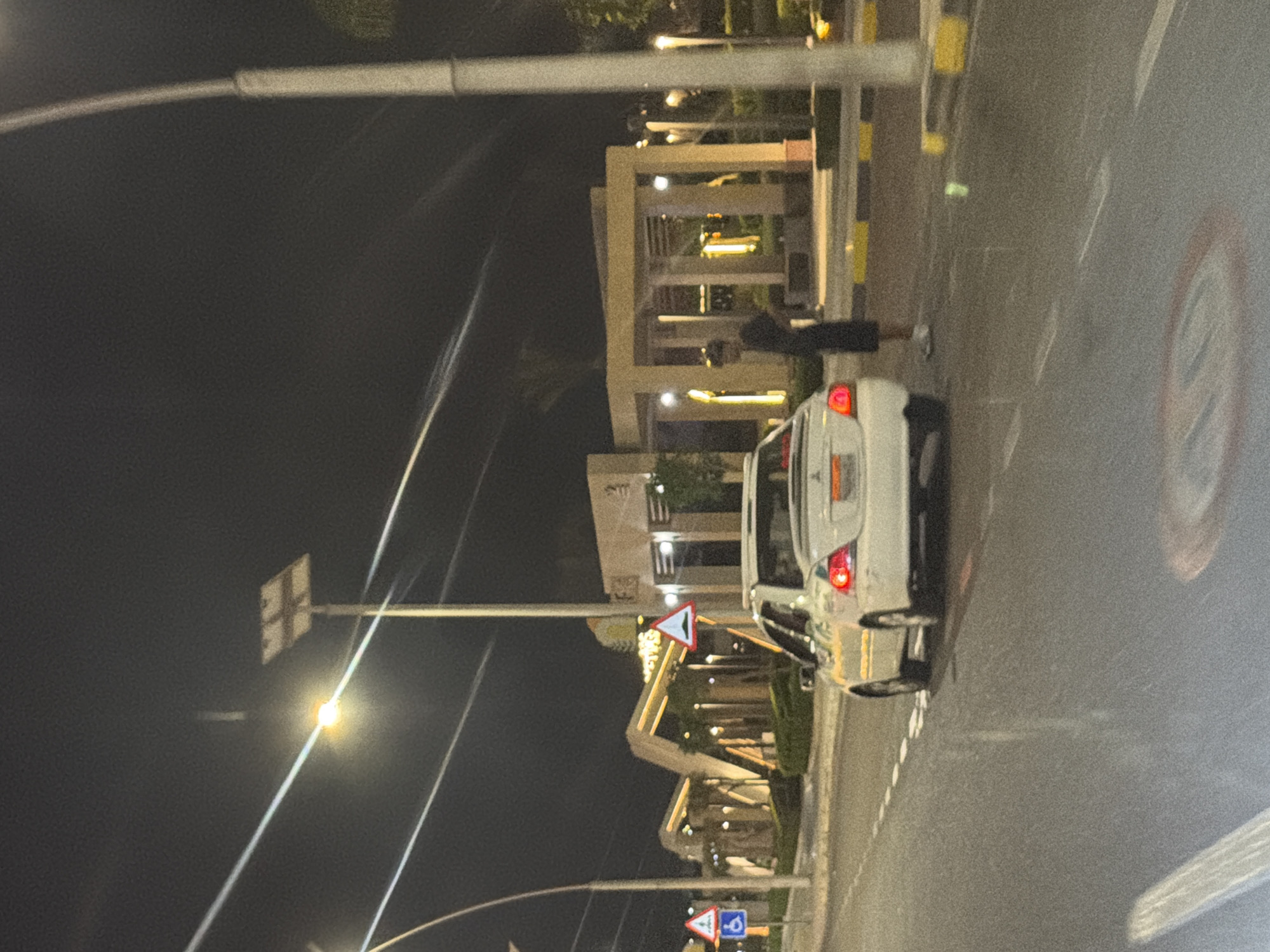 In the city of Sharm El Sheikh, Egypt, at night, a white sedan is parked on the right side of a paved road. A lone figure, a man wearing a dark shirt and shorts, stands on the sidewalk beside the car, facing away from it. The background features a modern architectural structure with illuminated elements, indicative of a commercial or recreational area. Streetlights and power lines are visible, contributing to the night-time urban ambiance. The road surface appears wet, reflecting the ambient light. Several traffic signs are present, including a blue sign with a white wheelchair symbol and a triangular sign with a green hill symbol.