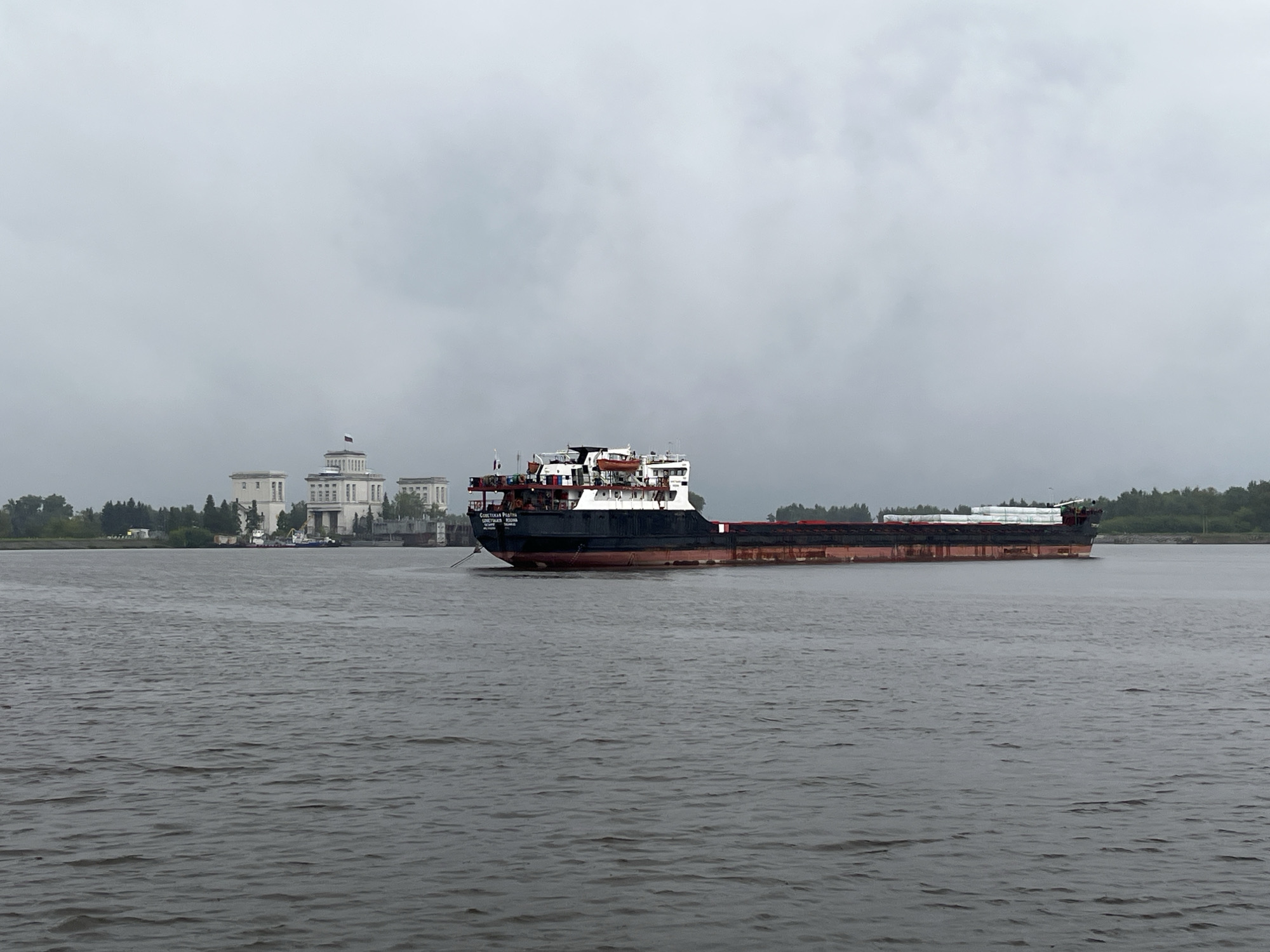 A wide-angle, exterior, eye-level, static shot shows a grey, overcast sky over a dark, choppy body of water. In the distance, on the left side of the frame, a large, white, neoclassical building with columns and a dome topped with a flag is visible. To the right of the building, a large cargo ship with a black hull and a red waterline is sailing on the water. The ship has a white superstructure with several levels and a crane. The ship is partially obscured by the building and the water. The shoreline behind the building is lined with trees. The overall atmosphere is muted and somber due to the overcast sky.