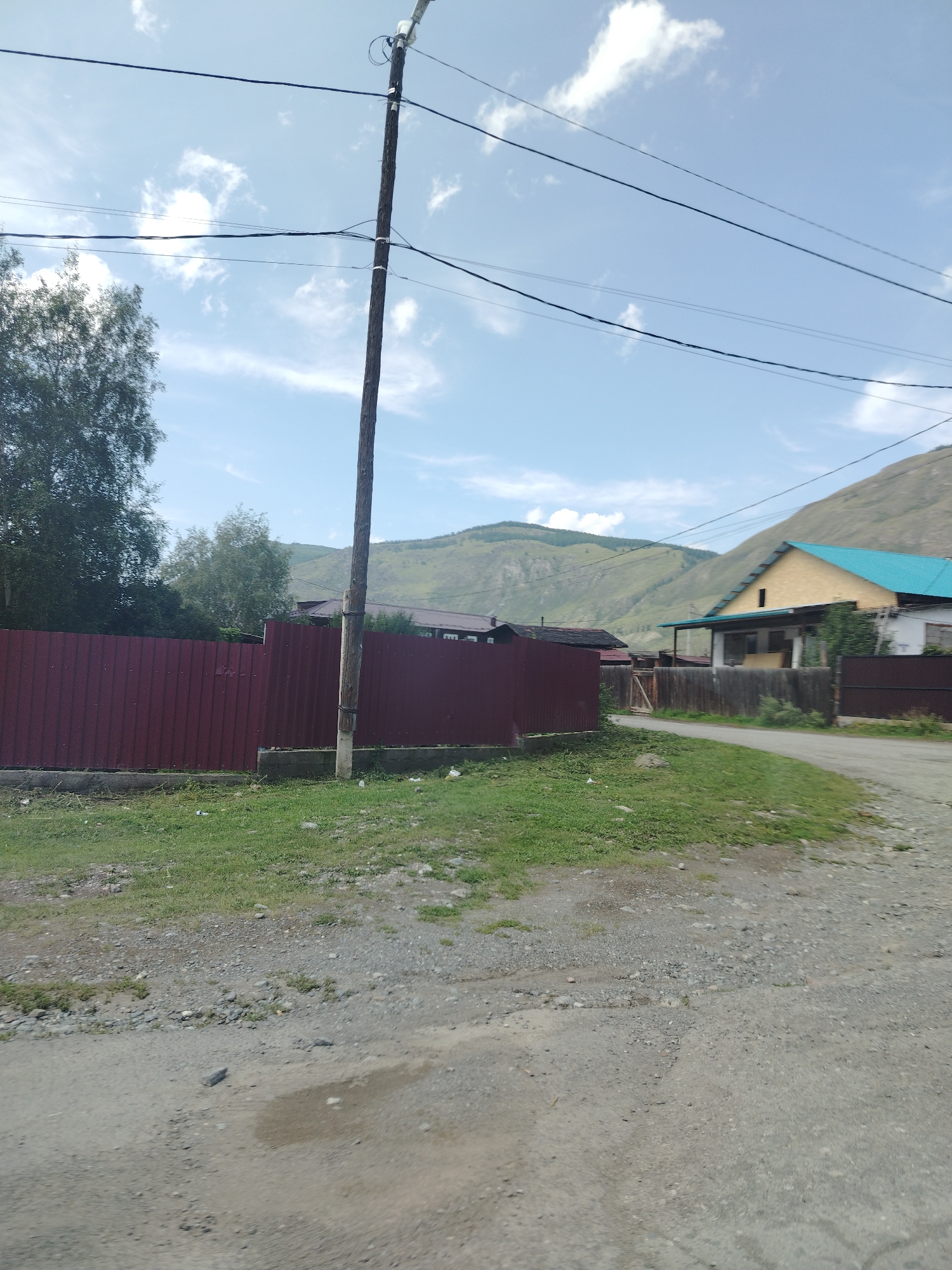 The image captures a street scene in Aktash, Russia, under a partly cloudy sky. A wooden utility pole with multiple wires runs vertically through the center of the frame, supporting overhead electrical or telecommunication lines that stretch across the sky. To the left, a dense green tree stands tall. In the middle ground, a long, dark red corrugated metal fence runs horizontally, bordering a patch of green grass. Beyond the fence, a residential area is visible, featuring houses with yellow and blue roofs, wooden fences, and some vegetation. The foreground consists of a gravel and dirt road with visible patches of mud and stones. In the background, rolling hills with sparse vegetation rise under the sky, indicating a rural or semi-rural setting within Aktash. There are no people or animals present in the image.