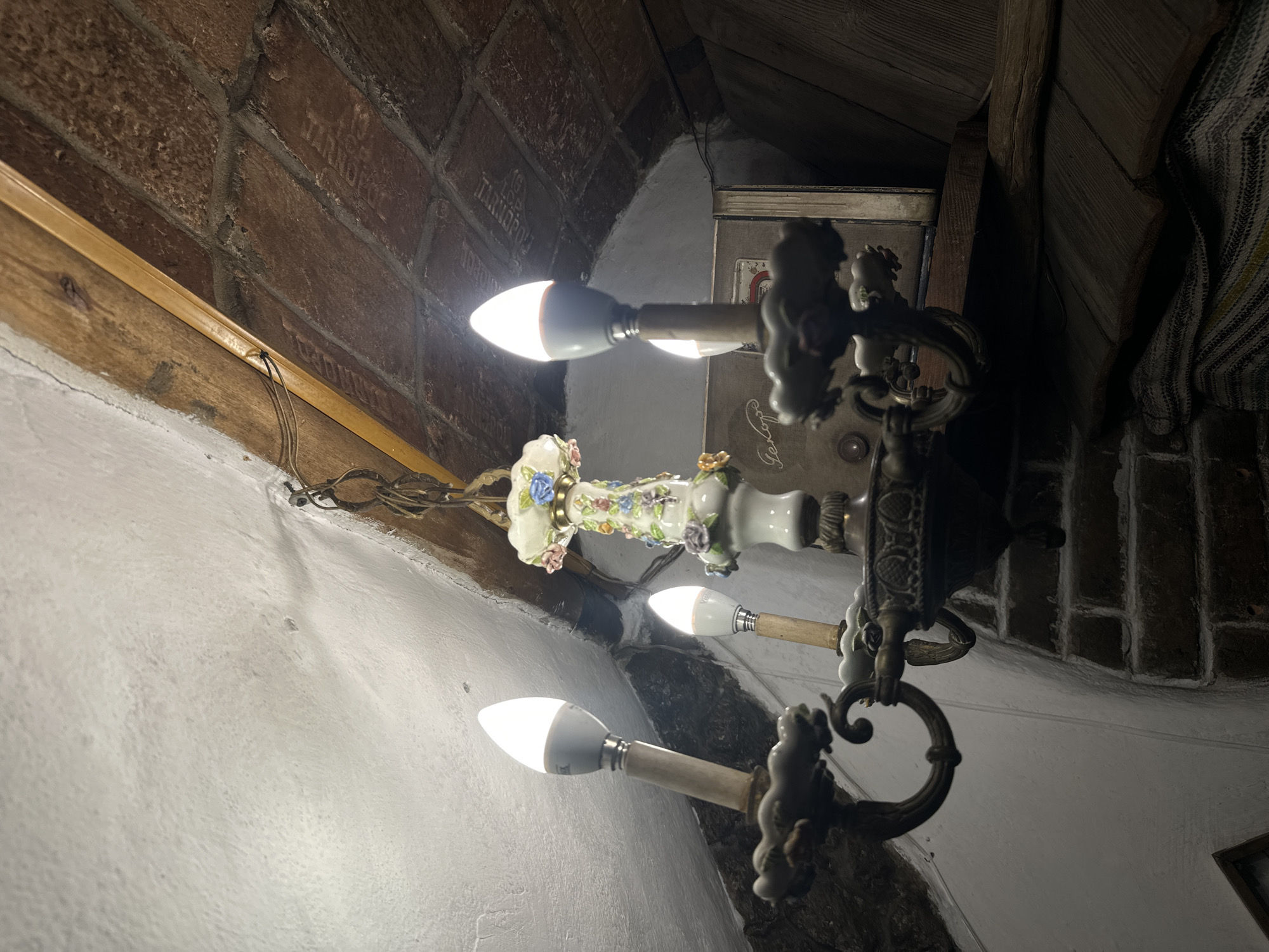 The image shows a close-up of a vintage chandelier hanging from a ceiling in what appears to be an interior space. The chandelier has a central decorative ceramic piece with floral motifs, from which metal arms extend to hold three candle-shaped LED bulbs. The ceiling is a mix of textures, with a white, possibly stuccoed surface on the left, and exposed brick and wooden beams on the right, suggesting an older building. A portion of what looks like a vintage radio or an old book with visible lettering can be seen on a shelf in the background. The lighting suggests a warm, ambient environment. The scene is static and does not contain any people or visible actions. No specific location in Ternopil, Ukraine is identifiable from the image itself.