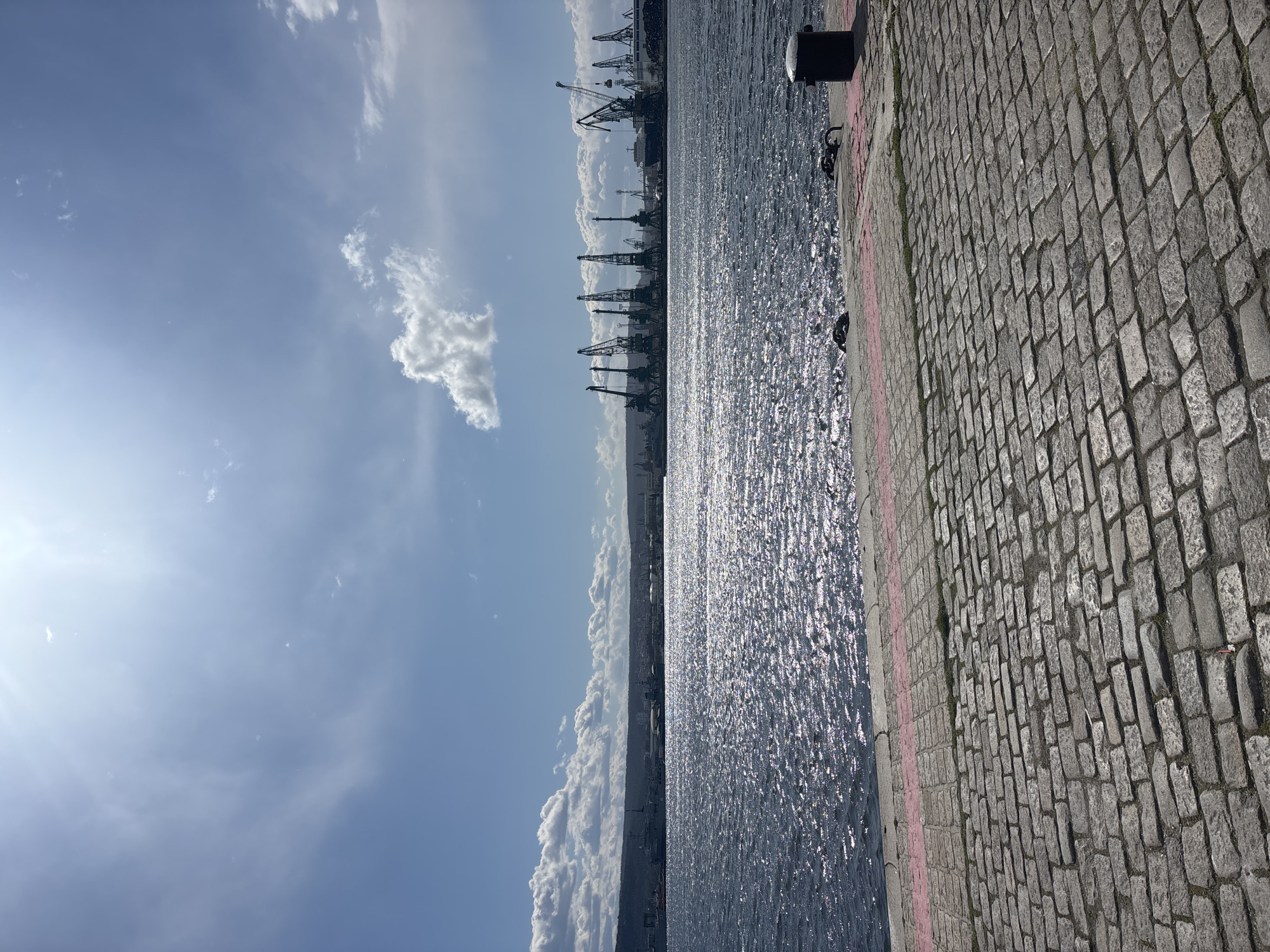 The image shows a waterfront scene in Varna, Bulgaria. The right side of the image features a stone-paved walkway with a distinct pink line marking its edge next to the water. A bollard is also visible. The water reflects sunlight, creating a sparkling effect. In the distance, a row of port cranes is visible along the coastline. The sky above is bright blue with scattered clouds. The image appears to be taken during the day.