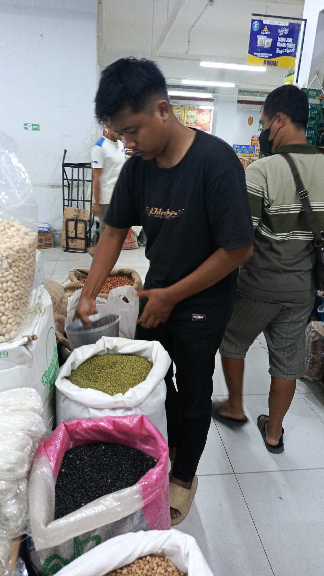 The image depicts an indoor market scene, likely a grocery store or traditional market, in Ponorogo, Indonesia. In the foreground, a young man wearing a black t-shirt and black pants is shown reaching into a sack of green beans with a metal scoop. He is focused on the task, with his head bowed. Behind him and to his right, another man wearing a striped t-shirt and shorts, with a black mask covering his lower face, stands looking away from the camera. Various large sacks of grains and beans are visible in the scene, including a large bag of peanuts to the left, a sack of green beans in the center, and a sack of black beans in the foreground with a pink trim. Another sack of what appears to be peanuts or a similar nut is visible behind the green beans. The floor is tiled, and overhead lighting illuminates the space. A metal rack is visible in the background to the left, and promotional banners are hanging on the wall further in the distance. The environment suggests a commercial setting where bulk food items are sold.