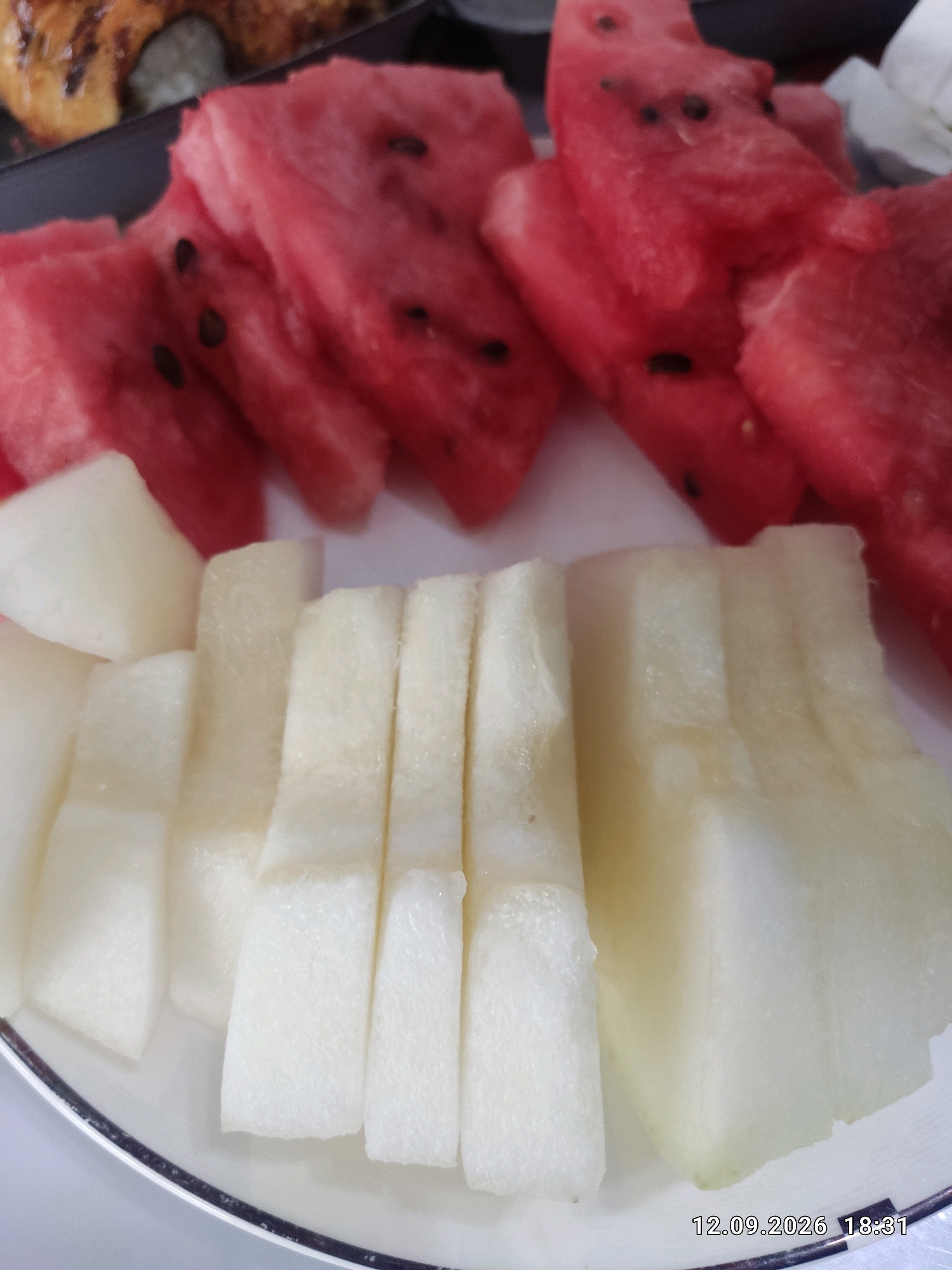 A close-up, top-down view of sliced fruits on a white plate shows several pieces of watermelon with seeds, and several pieces of pale yellow fruit, possibly honeydew melon. The fruits are arranged in two distinct sections on the plate, with the watermelon slices towards the top and the pale yellow fruit slices below them. In the upper left corner, partially visible, is a dark plate containing what appears to be a pastry or sushi roll. A timestamp, "12.09.2026 18:31," is superimposed on the bottom right of the image. The scene depicts food prepared for consumption, likely in a casual dining setting.