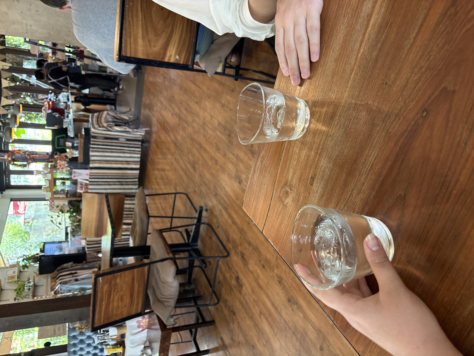 This is a medium-angle shot taken inside a cafe, looking down at a wooden table with two glasses of water on it. The scene suggests a casual gathering or meal. The lighting is soft and diffused, indicating it might be daytime, possibly late morning or early afternoon.
In the foreground, a person's hand is visible, holding one of the glasses. The glass contains water and ice cubes. The table surface is a warm, polished wood with visible grain patterns. In the background, the cafe's interior is visible, with glimpses of other tables, chairs, shelves stocked with items, and large windows offering a view of greenery outside. The decor appears eclectic, with elements of natural wood, textiles, and plants, suggesting a relaxed and possibly bohemian atmosphere.
The focus is primarily on the table and the glasses of water, creating an intimate feel. The presence of hands suggests human activity and interaction within the space. The overall mood is calm and convivial. The location context of Chiang Mai City Municipality, Thailand, might imply a warm climate, which would explain the presence of ice in the water. No visible text is apparent in the image.