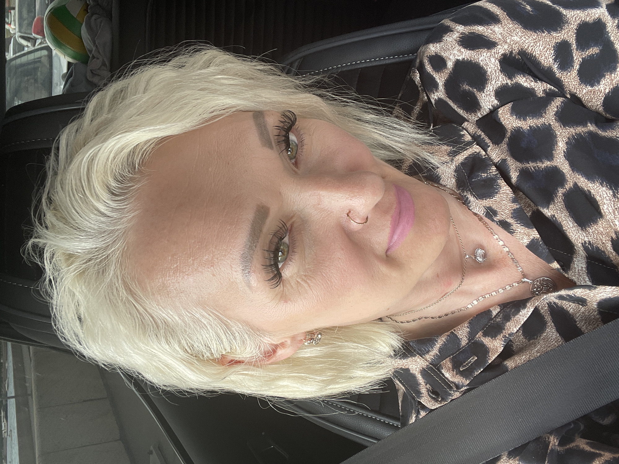 A woman with blonde hair and green eyes is sitting in the driver's seat of a car. She is wearing a leopard print shirt and two necklaces. Her hair is styled in loose waves and she is wearing makeup, including mascara, eyeliner, and pink lipstick. She has a nose ring and earrings. The interior of the car is visible, including a black seat and a seatbelt. In the background, other cars are visible, as well as what appears to be a body of water and buildings. The lighting suggests it is daytime.