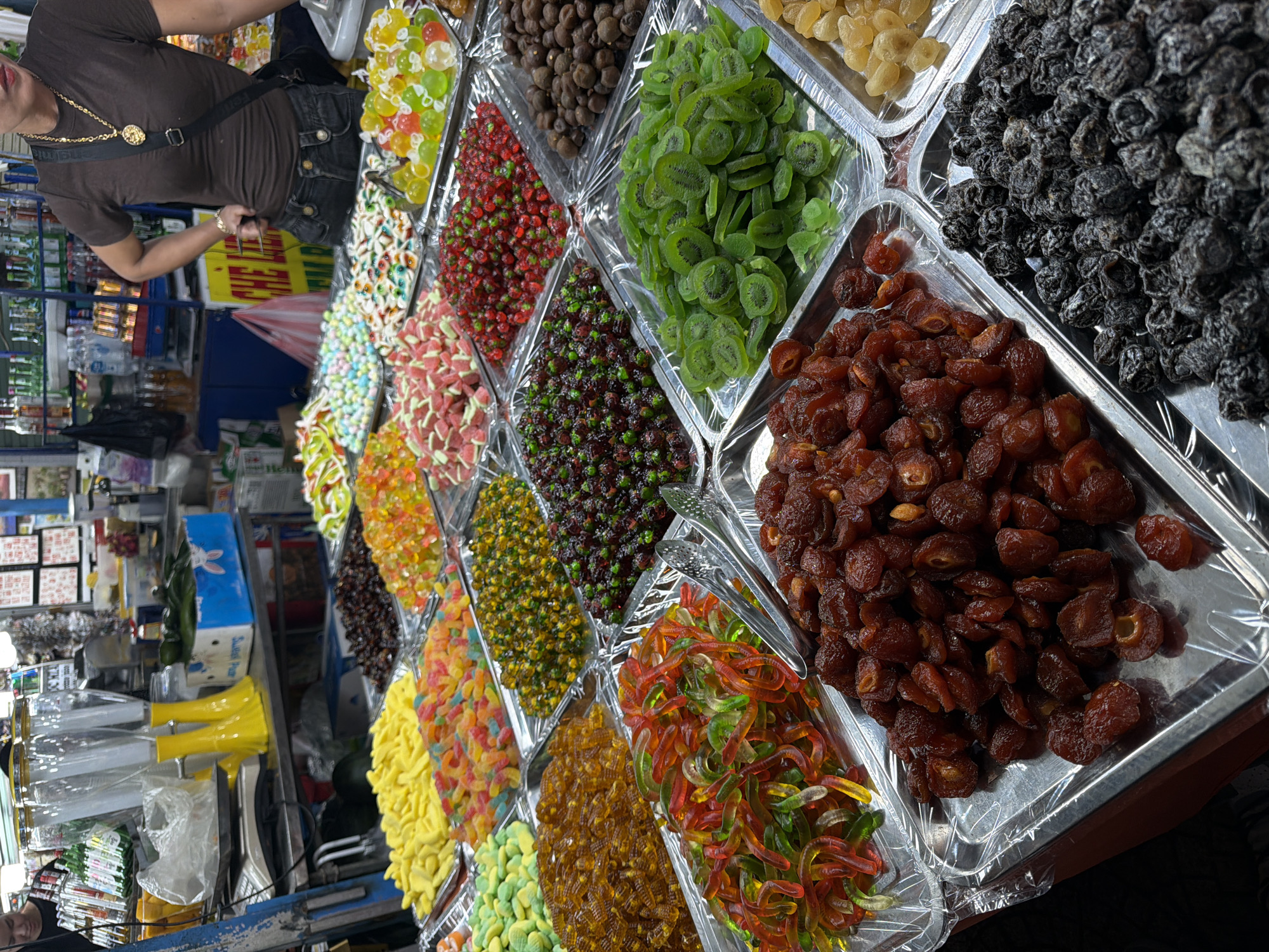 The image shows a street food stall in Đà Nẵng, Vietnam. Various types of candies and dried fruits are displayed in metallic trays, arranged in rows. The trays are covered with clear plastic wrap. From left to right, there are gummy worms, assorted gummy candies in different colors, dried fruits, sliced dried kiwi, and dried plums. A person wearing a brown shirt and a gold necklace stands in the background, near a section of the stall displaying assorted goods on shelves.