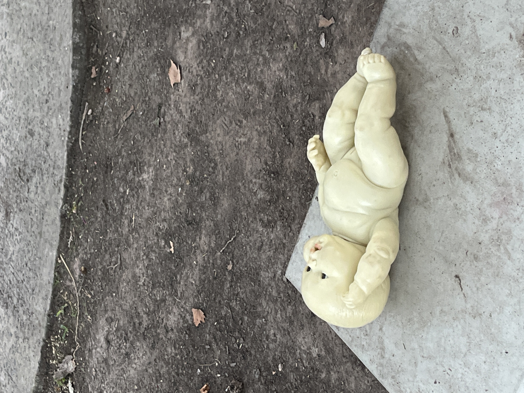 The image depicts a light yellow baby doll lying upside down on the ground in an outdoor setting in Olivos, Argentina. The doll, possibly made of plastic or ceramic, appears somewhat aged and dirty, with its limbs bent in a static position. The scene is divided by two distinct ground surfaces: the left side features dark, loose earth containing small debris such as dried leaves and twigs, while the right side, where the doll is positioned, consists of a lighter, mottled grey surface, likely concrete or pavement. Natural, diffused light illuminates the scene, suggesting daytime with overcast conditions or indirect sunlight. There is no specific activity taking place, and the image conveys a sense of quiet stillness or abandonment. No visible text is present.