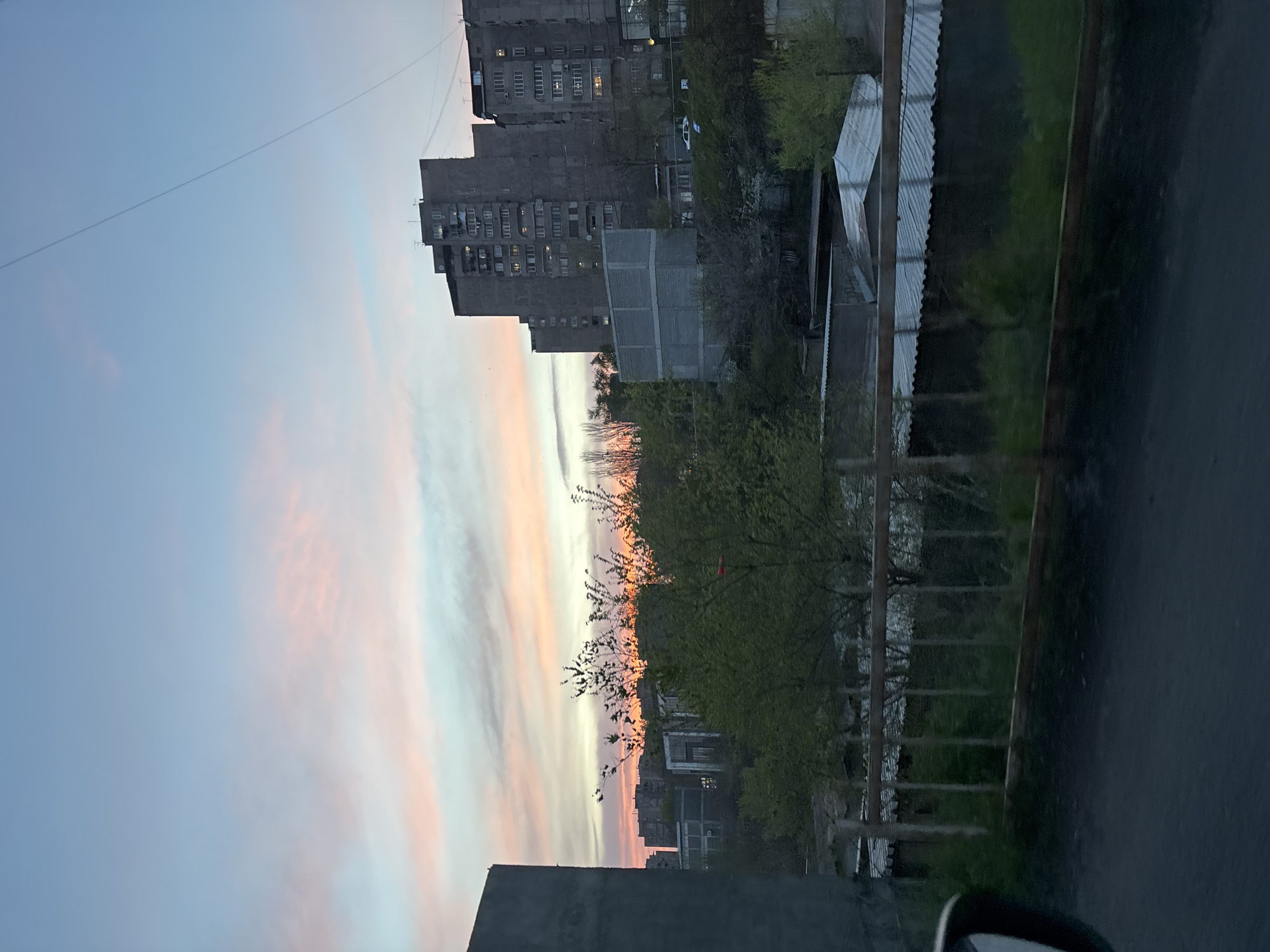 The image depicts an urban or semi-urban landscape in Yerevan, Armenia, captured during either sunrise or sunset. The scene is dominated by a dramatic sky and utilitarian architecture.
The sky, occupying the upper-left portion of the frame (when correctly oriented), transitions from a soft light blue at the top to striking horizontal streaks of pink, orange, and pale yellow clouds across the middle, indicating twilight. The overall illumination is dim, creating a serene atmosphere.
In the mid-ground and right side, a large, multi-story concrete residential building, typical of Soviet-era construction, stands prominently. Several windows emit a warm light, suggesting occupied apartments. Below and in front of this main structure are other concrete buildings and smaller structures with corrugated metal roofs, partially obscured by trees.
Numerous trees are scattered throughout the scene. Some appear to be budding or have sparse leaves, while others boast fuller green foliage, providing a natural element against the concrete backdrop. A distinct branch with fresh green leaves is silhouetted against the vibrant sky.
The extreme right edge of the image features a dark, asphalt-like surface, likely a road or path. A metal fence or railing runs parallel to this surface, separating it from the green areas and buildings. A thin wire or cable stretches diagonally across the upper sky. A faint, light-colored object, possibly a vehicle, is barely visible behind some trees in the background.
No people or specific activities are visible, presenting a quiet, contemplative snapshot of the urban environment at dawn or dusk. There is no visible text in the image.