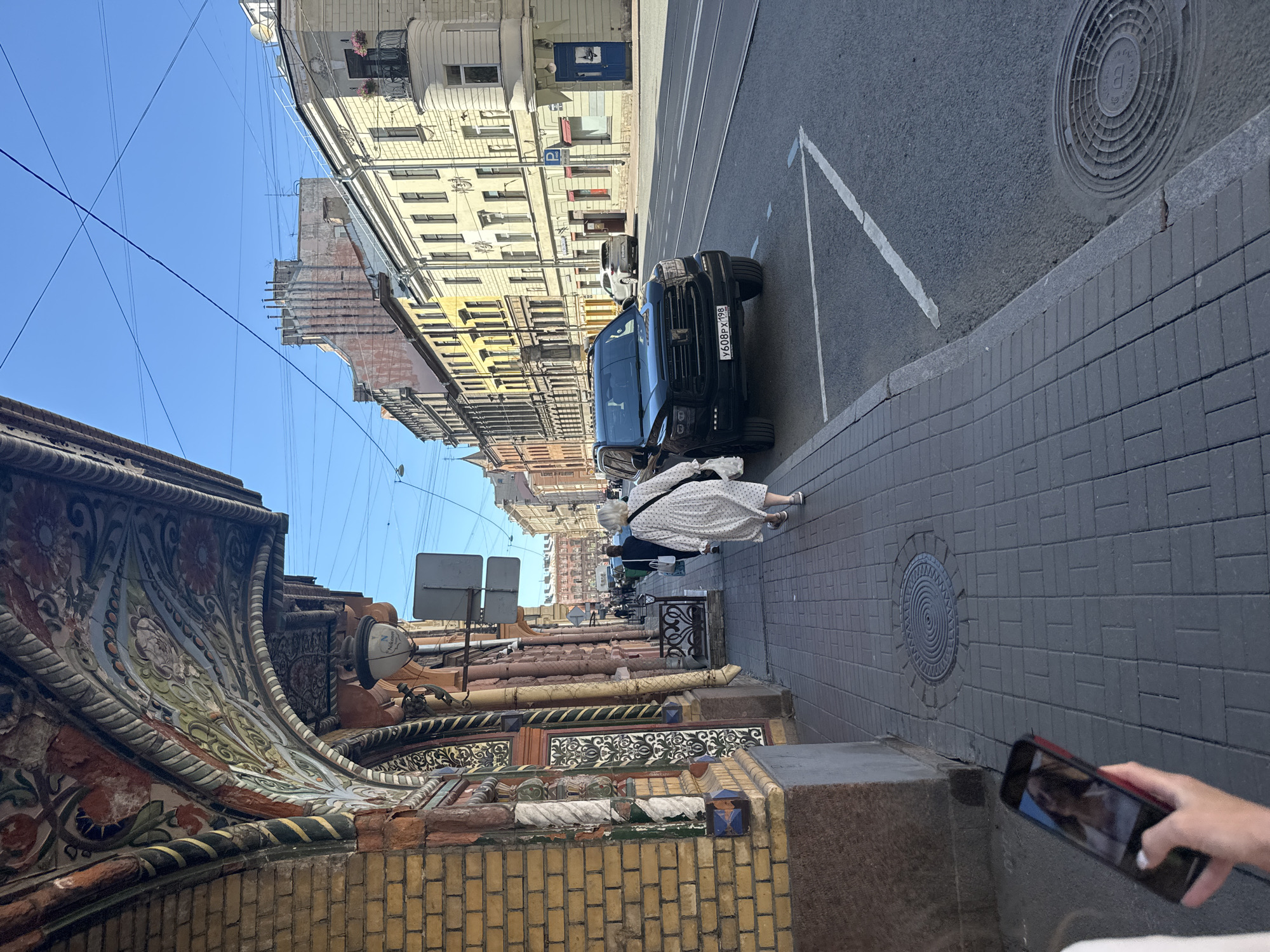 The image depicts a street scene in Saint Petersburg, Russia, on a sunny day. In the foreground, a person's hand holds a smartphone displaying a selfie. The sidewalk is paved with gray bricks, and a manhole cover is visible. To the left, a building features a yellow brick facade and an elaborately decorated arched entryway with colorful floral motifs. Further down the sidewalk, a woman in a white dress with a black polka-dot pattern walks away from the camera. A black SUV with a license plate "Y608PX 198" is parked on the street. The street is lined with buildings of various architectural styles and colors, including yellow and cream facades, with some displaying balconies. Overhead, a network of power lines crisscrosses the clear blue sky. In the distance, the street extends, with more buildings and a few other pedestrians visible.