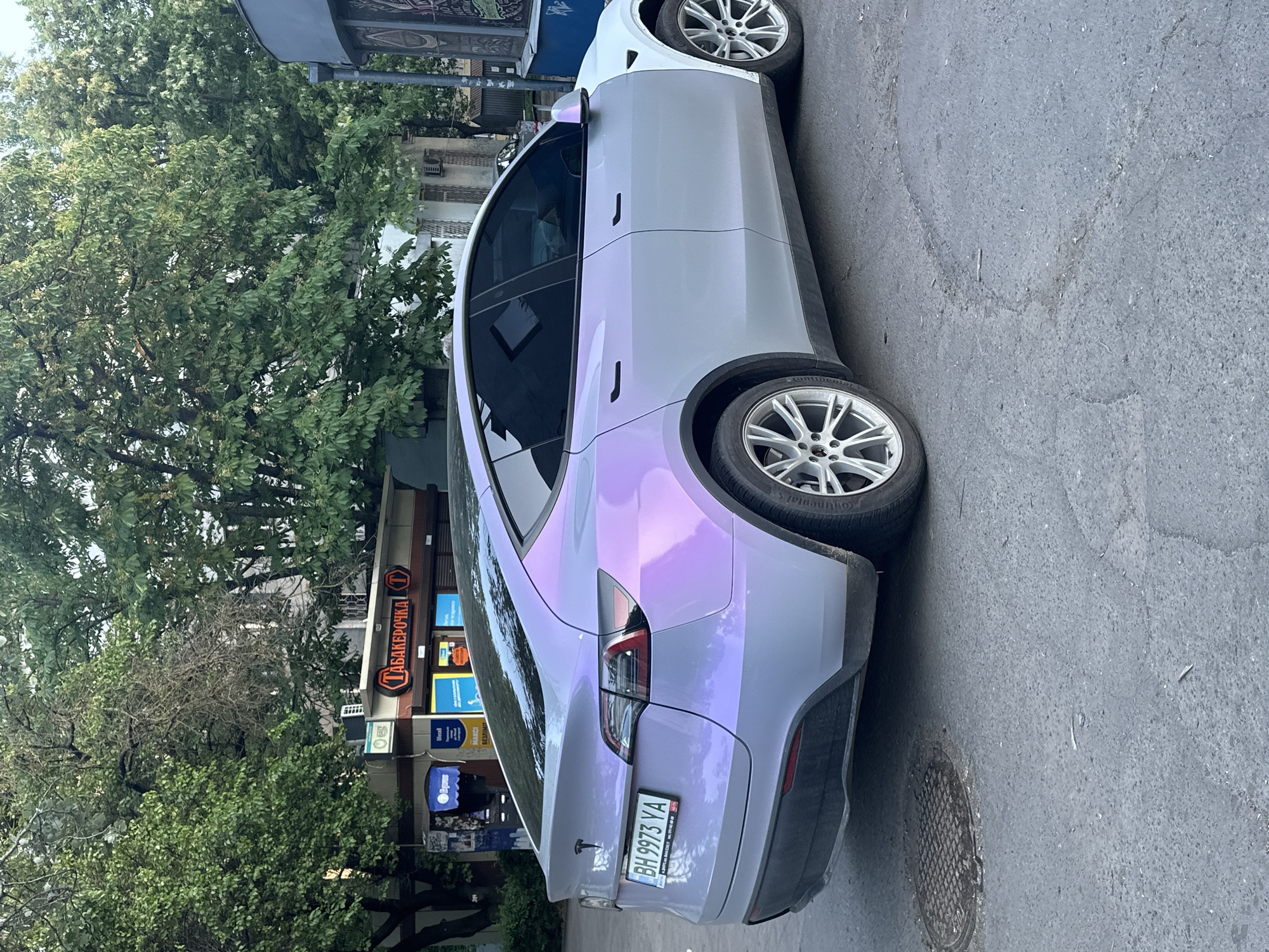 A Tesla Model Y with a two-tone color scheme of white and purple is parked on the street in the city of Unknown, Unknown. The car has a license plate with the number "BH 9973 YA." In the background, there are trees and a building with a sign that reads "ТАБАКЕРОЧКА" in Cyrillic.
