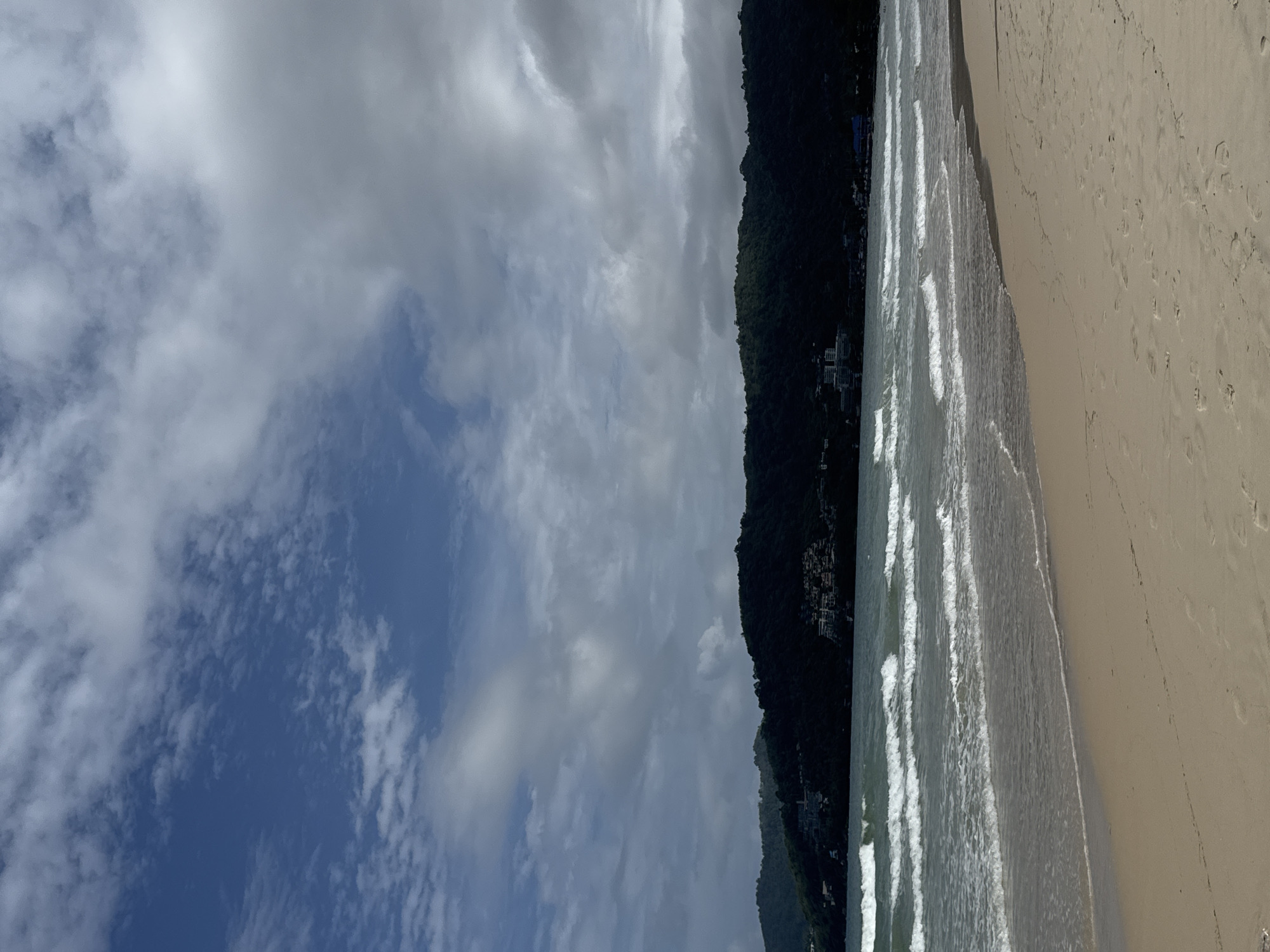 The image depicts a wide, sandy beach in Patong, Thailand, with the Andaman Sea in the background. Gentle waves are rolling onto the shore, creating white foam. In the distance, lush green hills rise up, dotted with buildings, suggesting a developed coastal area. The sky is partly cloudy, with patches of blue visible between the larger grey and white clouds. The sand is light brown and shows some footprints, indicating recent human presence. The overall scene is serene and natural, with hints of a populated tropical destination.