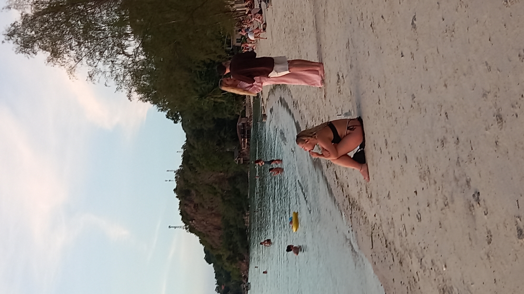 The image depicts a beach scene in ตำบลเกาะพะงัน, Thailand. In the foreground, a woman in a black bikini is seated on the sand, facing away from the camera, with her legs crossed and her hand near her mouth. Further back, a couple stands close together on the sand, the woman in a long pink dress and the man in a brown shirt and white shorts. To their left, several people are in the shallow water. Some are swimming, while one person appears to be using a yellow inflatable ring. The water is calm and reflects the sky. In the background, a lush, tree-covered hill rises from the water, with some buildings visible amongst the trees. The sky is a pale blue with some wispy white clouds. The overall atmosphere suggests a relaxed, tropical setting.