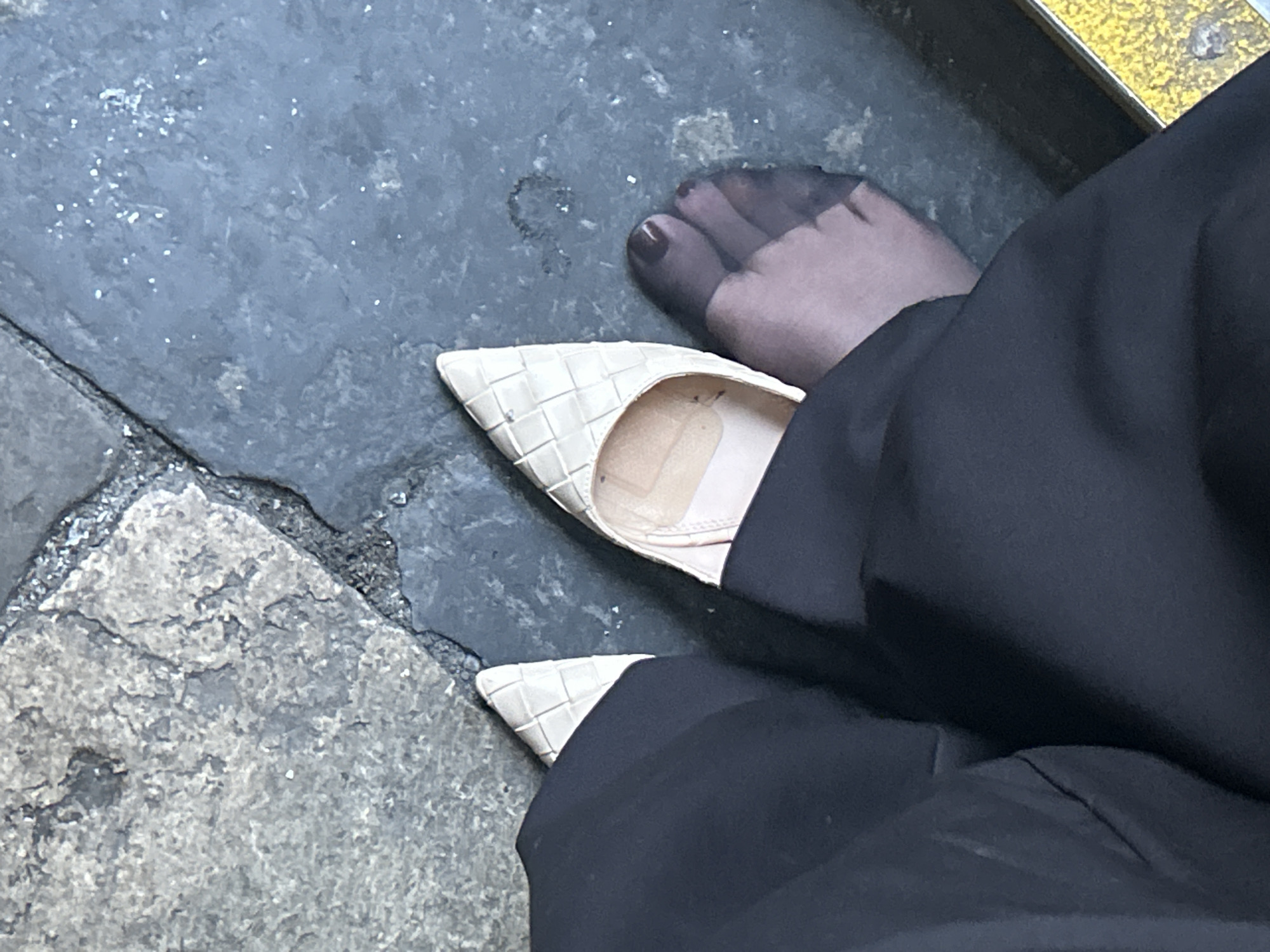 The image presents a close-up, high-angle view of a person's lower leg and foot, partially within a shoe, resting on outdoor pavement. The foot is encased in sheer dark hosiery, revealing toes adorned with dark red nail polish. The foot is partially slipped out of a light-colored, likely beige or off-white, pointed-toe flat shoe. The shoe features a distinctive woven or basketweave texture across its surface. Dark, loose-fitting trousers or pants cover the upper part of the person's leg and partially obscure the shoe.
The ground consists of dark grey, textured pavement, possibly asphalt or rough stone, displaying visible cracks and some lighter, worn areas. Scattered small, shiny flecks, which could be glitter, moisture, or reflective particles, are visible on the darker surface. In the bottom-left corner of the frame, a lighter, rough-textured stone slab or flagstone is visible, indicating varying pavement types. A small section of a yellow, textured object with a metal bolt is visible in the top-right corner, possibly part of a kerb or street furniture.
The activity depicted is subtle, suggesting the person might be resting, adjusting their shoe, or having their foot slip out temporarily. The setting appears to be an urban environment, consistent with the provided location context of London, United Kingdom, though no specific landmarks are visible. The time of day and weather conditions are not discernible. There is no visible text in the image.