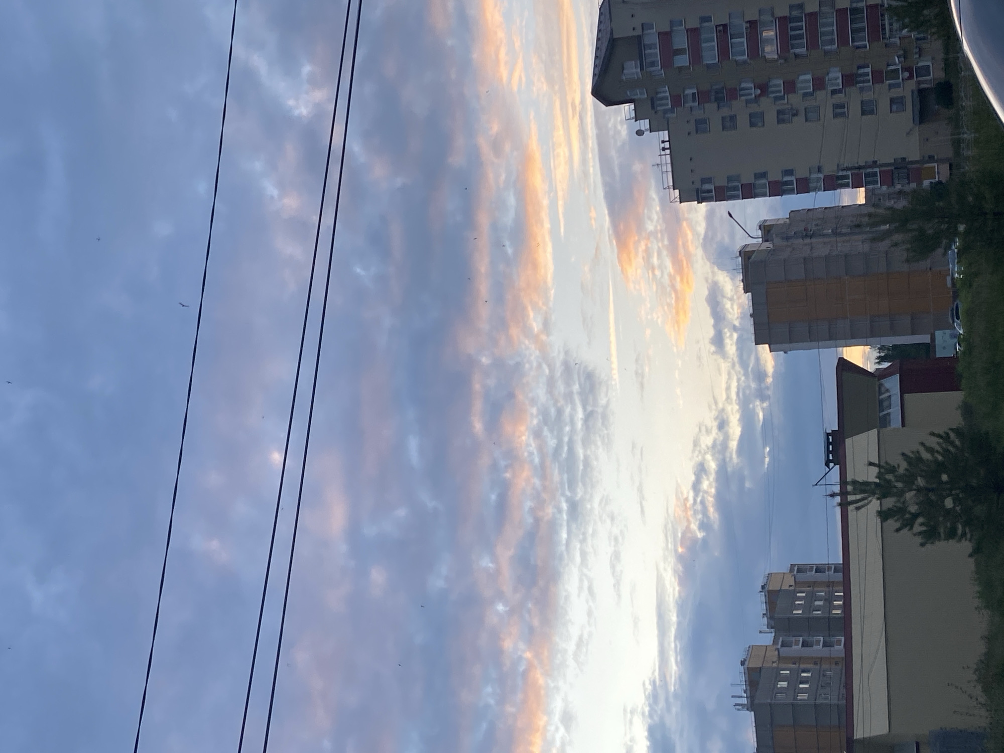 This is a medium-shot photograph of a city skyline at dusk. The sky is filled with dramatic clouds tinged with pink and orange, indicating the setting sun. Three power lines traverse the upper portion of the frame horizontally. In the foreground, several multi-story buildings are visible. On the far right, a tall, modern apartment building with red and white accents stands out. To its left, another building with yellow and gray facades is partially visible. In the mid-ground, a low-rise building with a red roof is present, and to its left, more apartment buildings are seen. A small evergreen tree stands in front of the red-roofed building. The overall scene depicts an urban environment under a colorful twilight sky.