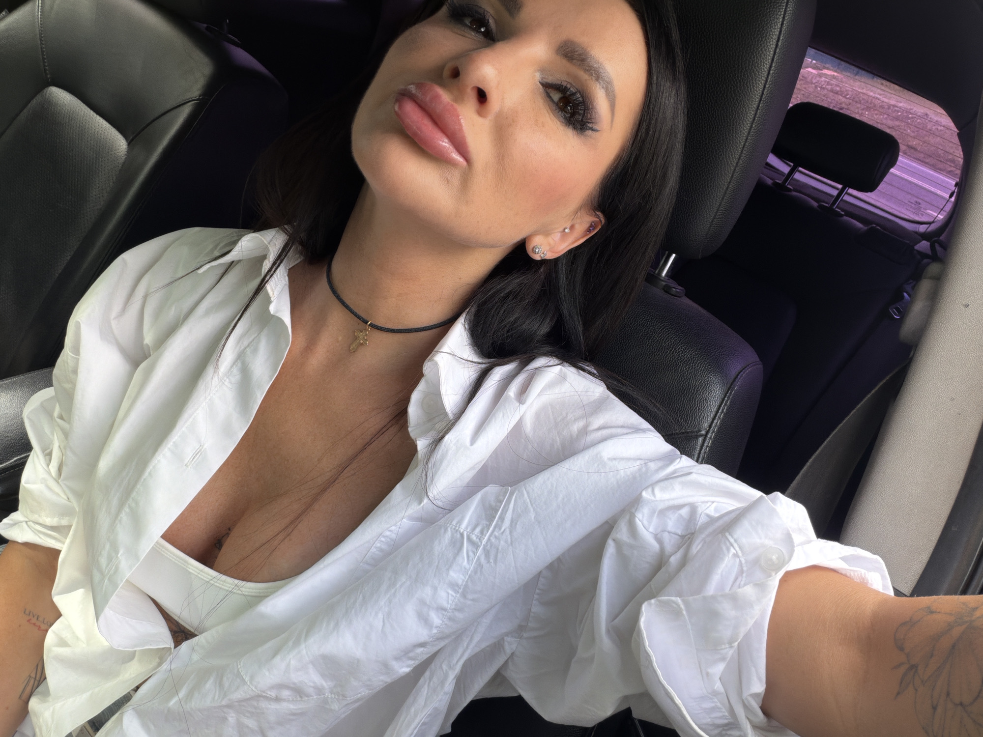 A woman is taking a selfie in a car. She is wearing a white button-down shirt, a white bralette, and a black choker necklace with a gold cross pendant. Her hair is dark and long, and she has dramatic eye makeup. She is wearing several earrings on her left ear. She has tattoos on her left arm, one of which has the text "LIVE LOUDLY." The interior of the car, upholstered in black leather, is visible. The lighting suggests it is daytime. The scene is likely in Novaya Usman', Russia, given the location context. The woman's expression appears confident and relaxed.