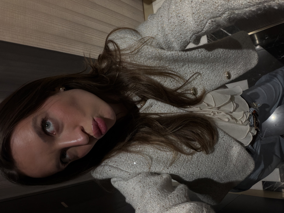 A woman is seen in a close-up shot from a slightly tilted, overhead perspective. She has light skin, brown hair, and is wearing a light-colored, textured jacket over a ruffled shirt. The jacket has shiny buttons. She is also wearing light blue pants. Her lips are pursed, and she is looking at the camera with blue eyes. The background is a mix of light and dark surfaces.