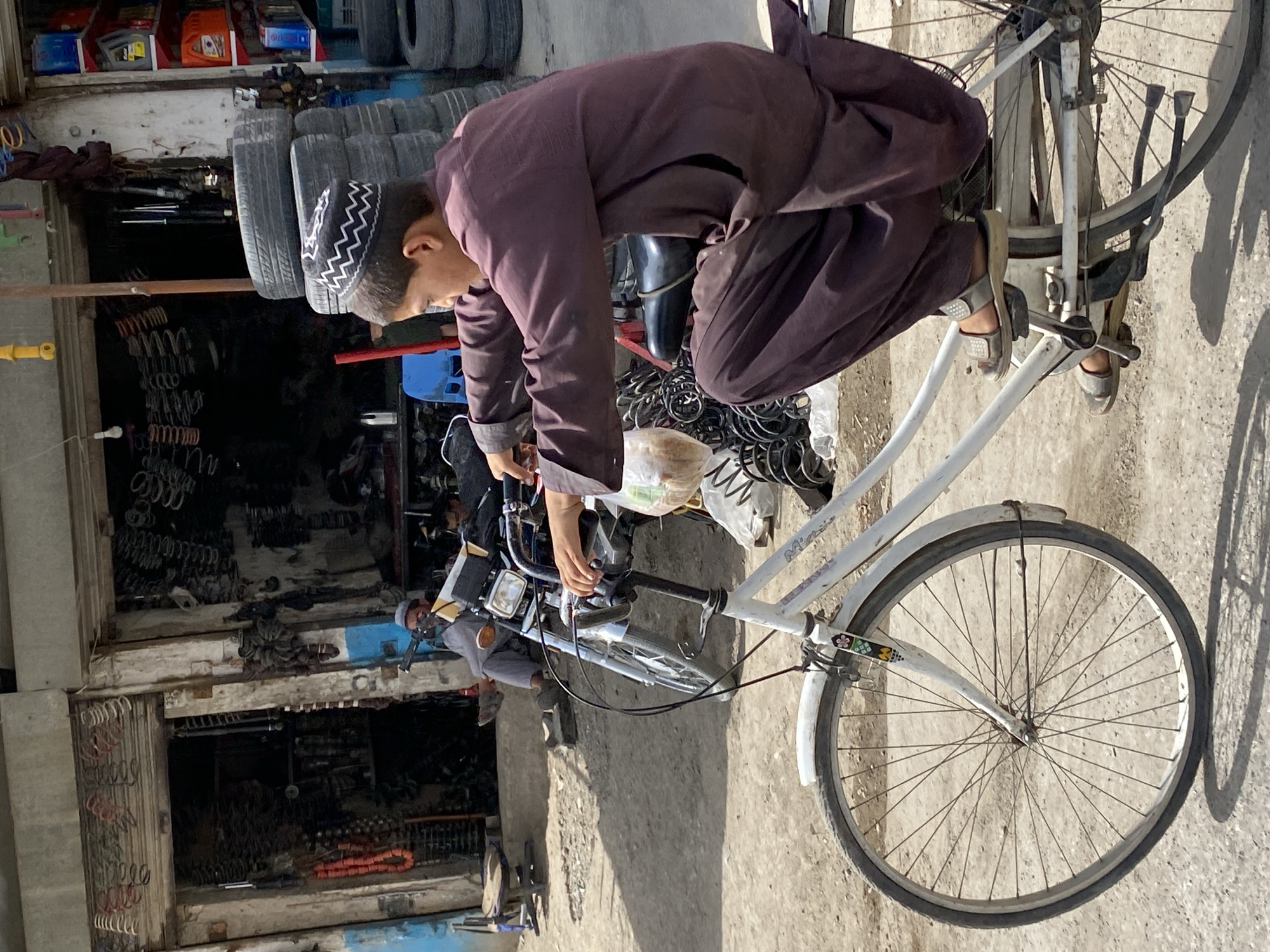 A young boy wearing a dark brown kameez and white, zig-zag patterned kufi is crouched on a white bicycle in the foreground, facing to the right. He is holding the handlebars with both hands and his feet are on the pedals. His face is turned away from the camera. The bicycle's front wheel is large and spoked. Behind the boy, a street scene unfolds. To the left, a store with an open roll-up door displays various metal coils and parts. A man in light gray clothing sits outside this store. To the right of the boy, a stack of tires is visible, along with a shop selling automotive parts. The ground is dusty and appears to be paved with rough concrete or dirt. The architecture suggests a Middle Eastern or South Asian urban environment. The overall impression is one of a busy marketplace or repair shop area in Kandahar, Afghanistan, on a sunny day.
