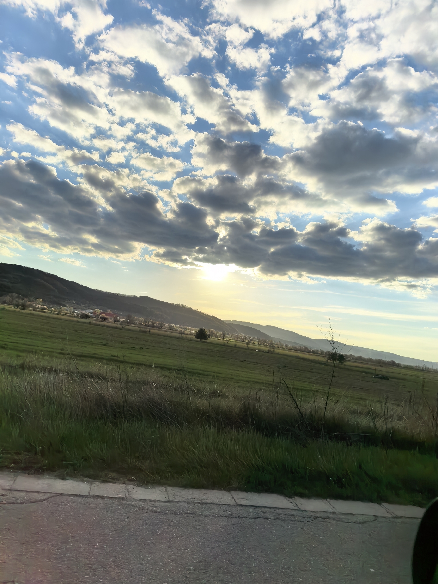 The image presents a wide view of a scenic rural landscape, likely captured during late afternoon or early morning. The sky occupies a significant portion of the frame, filled with an array of cumulus clouds. Some clouds are brightly illuminated with warm yellow and white tones by the sun, while others appear darker grey, interspersed with patches of clear blue sky. The sun itself is visible in the mid-right, peeking through or from behind the clouds, casting a soft, golden glow across the scene.
In the foreground, a strip of asphalt road or pavement with a concrete curb runs along the bottom. Immediately beyond the curb, there's a band of tall, dry grass transitioning into lush green fields that stretch into the midground. These fields gently slope upwards towards the distant hills. Scattered trees, some with light-colored blossoms, are visible across the fields and at the base of the hills, suggesting spring.
In the mid-left, a small cluster of houses with reddish roofs can be seen nestled at the foot of a prominent, dark green hill, hinting at a village or settlement. Further in the background, a range of rolling hills or mountains extends across the horizon, appearing hazy in the soft light. A few bare branches or a sparse bush are visible in the right foreground. An indistinct dark shape, possibly part of a vehicle, is present in the very bottom right corner.
There are no people or specific activities taking place. The scene conveys a peaceful and natural ambiance. No visible text is present in the image.