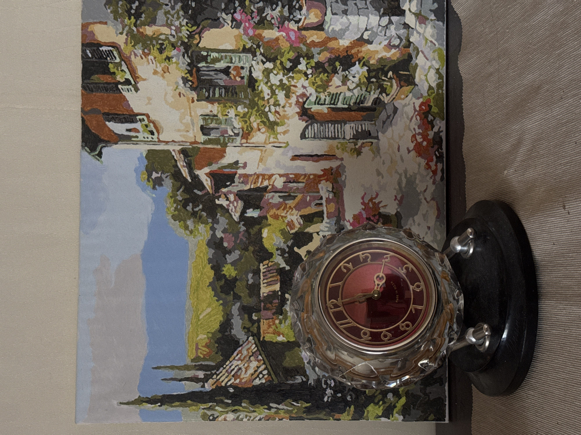 A framed painting of a quaint European cityscape, with buildings, greenery, and a blue sky, sits next to a vintage, crystal-encased table clock with a red face. The clock is mounted on a black base with chrome accents. The scene is photographed on a beige-toned surface. The location of Kazan, Russia is not definitively identifiable from this image.