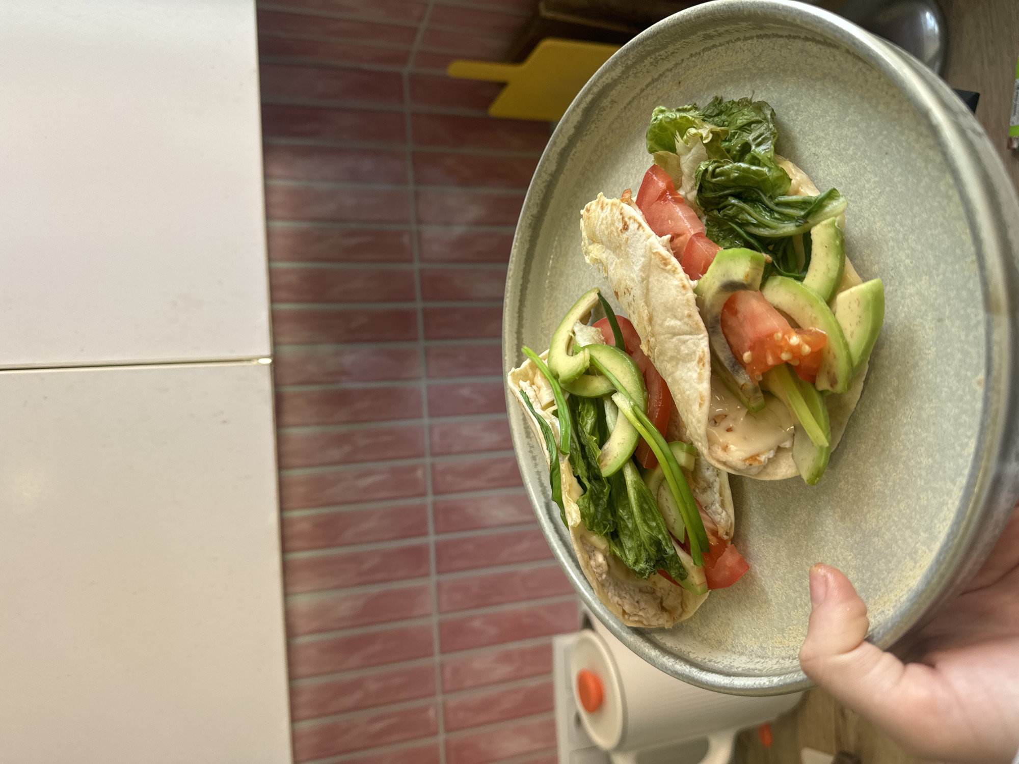 A person holds a grey plate containing two tortilla wraps. The wraps are filled with lettuce, tomato, avocado, and white sauce. The background shows a white cabinet, a red tiled wall, and a yellow kitchen utensil. The location and city details are not discernible from the image.