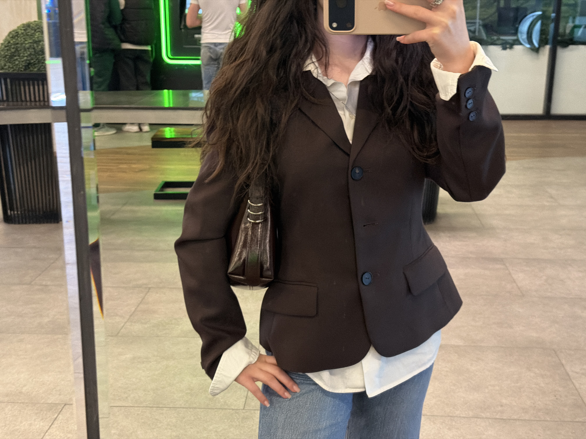 A person wearing a brown blazer, white collared shirt, and blue jeans is standing indoors, possibly in a commercial space with reflective flooring. The person is holding a mobile phone, seemingly taking a photo or a selfie. A small brown purse is slung over the person's shoulder. In the background, there are people and what appears to be a neon-lit feature. The location cannot be definitively determined as Yerevan, Armenia based on the visual information provided.