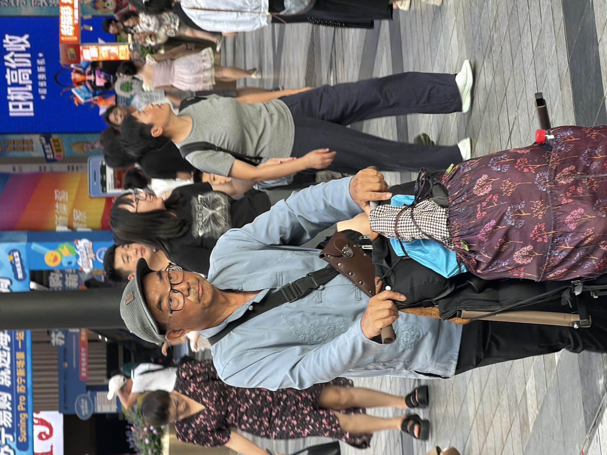 In Yuzhong District, China, a man wearing a light blue collared shirt, dark pants, and a gray cap pushes a shopping cart loaded with bags. He is wearing glasses and has a serious expression. He is walking on a paved sidewalk crowded with pedestrians, including women in dresses and other individuals. The background shows a busy urban environment with shops and advertisements. The man is the central figure, focusing on his task of moving the cart.