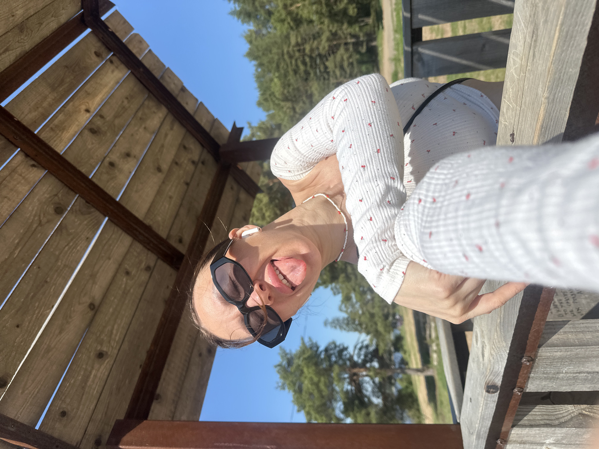 A woman is taking a selfie in a park-like setting, possibly in Votkinsk, Russia. She is wearing a white long-sleeved shirt with a cherry print, black sunglasses, and a beaded necklace. She has an earbud in her left ear and is sticking her tongue out. The photo is taken from a low angle, looking up at her under a wooden structure. Trees and blue sky are visible in the background. A weathered wooden railing is present in the foreground.