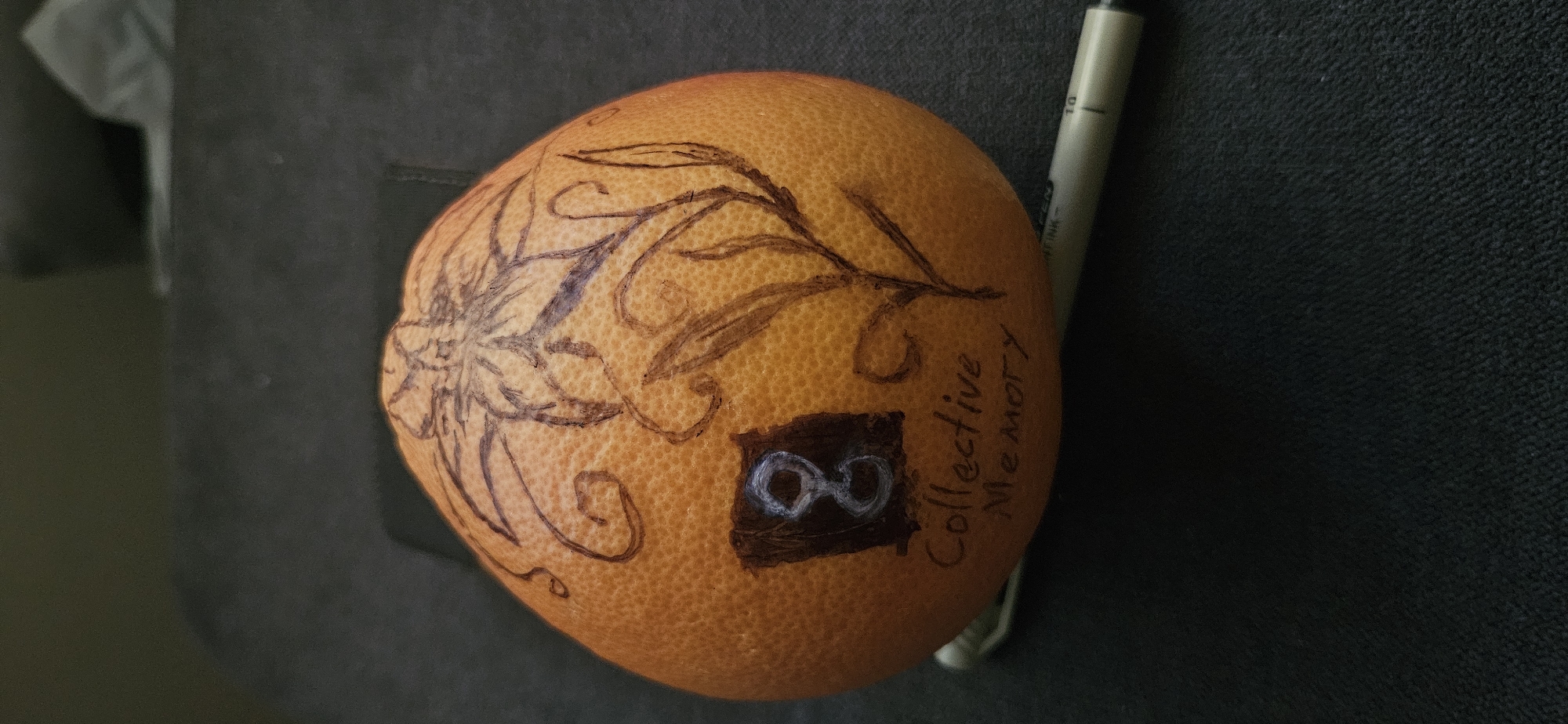 An orange grapefruit sits on a dark gray surface. The grapefruit is decorated with dark ink drawings of a stylized flower with leaves and branches, as well as a dark square containing a white infinity symbol. Next to the square are the words "Collective Memory" written in dark ink. A black and silver pen rests beside the grapefruit. The scene is not explicitly identifiable as being in Plovdiv, Bulgaria.
