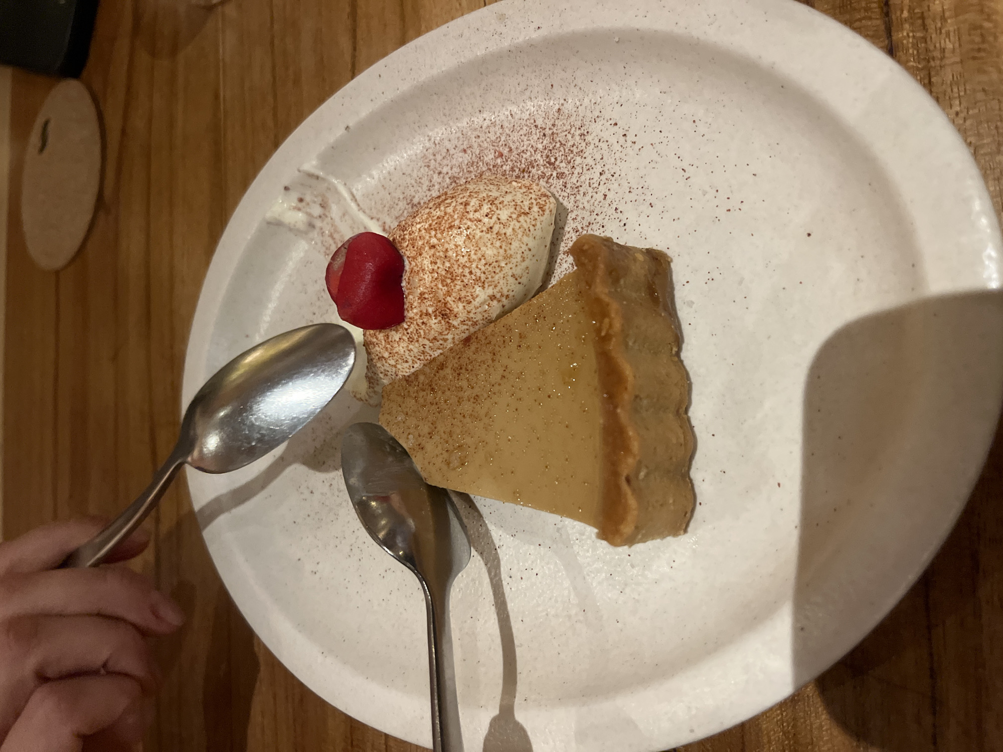 The scene is a close-up, overhead shot of a dessert on a white plate, likely taken in a restaurant or dining environment in Kuta Selatan, Indonesia. The dessert consists of a slice of golden-brown flan-like pudding, a dollop of whipped cream dusted with a reddish-brown spice (possibly cinnamon or cocoa), and a red berry or fruit slice. Two metal spoons lie on the plate, positioned near the dessert. The plate rests on a wooden surface. A human hand partially appears in the lower-left corner of the frame, holding one of the spoons. A circular coaster is also visible in the upper left of the image.