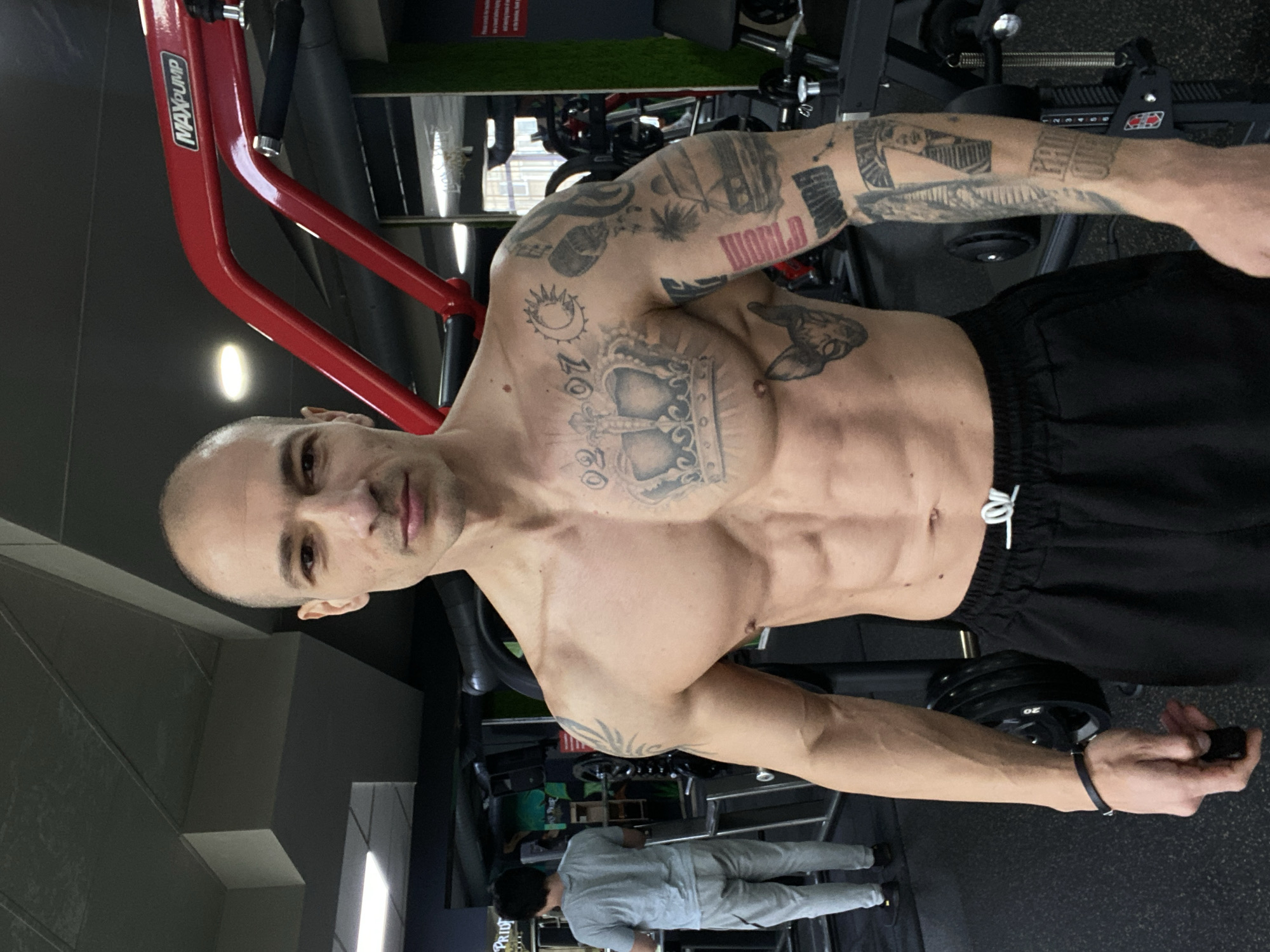 A man with a shaved head and muscular physique is standing in a gym. He is shirtless, wearing black athletic shorts with a white drawstring. He has numerous tattoos covering his torso and arms. One tattoo prominently features a crown, while another reads "WORLD". He is holding a small black object in his right hand.
The gym appears to be well-equipped with various weight machines. A red "MAXPUMP" machine is partially visible in the background. In the distance, another individual wearing a grey tracksuit is walking in the gym.
The location is identified as Pleven, Bulgaria.