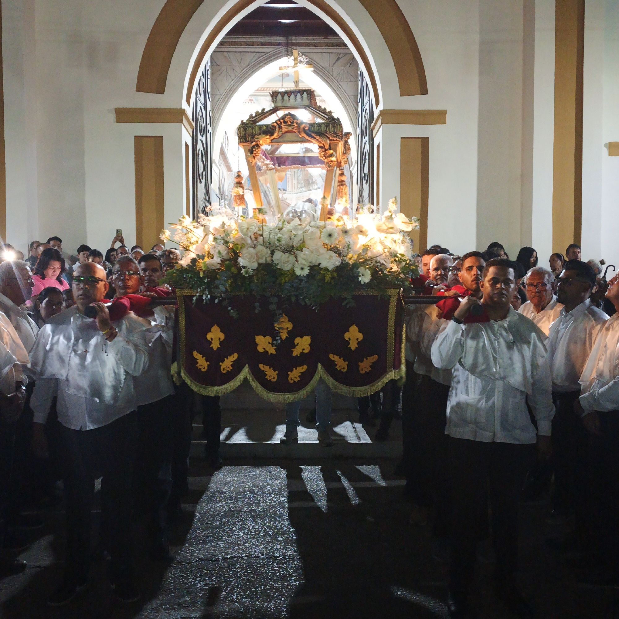 A procession in Tinaquillo, Venezuela, showcases men carrying a religious float adorned with white flowers and a burgundy cloth with gold fleur-de-lis designs. The float is elaborately decorated and illuminated. The men wear white shirts and dark trousers. They proceed through a building with arched doorways and onlookers stand by, some holding cellphones to capture the event. The lighting is mixed, with bright spots highlighting the float and darker areas near the ground.