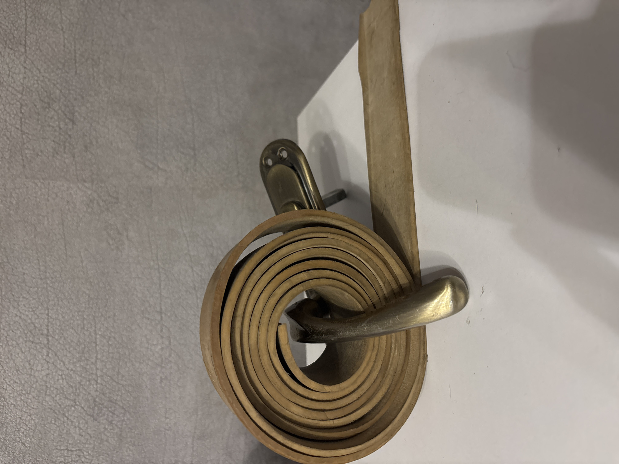The image shows a close-up view of a coiled leather belt, its light brown color and textured surface clearly visible. The belt is wrapped around a brass-colored metal object that appears to be a door handle or a similar piece of hardware. This metal object has a curved, ergonomic shape and two small holes near the top, suggesting it's meant to be attached to something.
The coiled belt is the prominent subject, filling a significant portion of the frame. The leather has a matte finish, and the edges of the belt are rounded. The way the belt is coiled creates a visually appealing spiral pattern. The metal hardware it's wrapped around provides a metallic contrast to the organic texture of the leather.
The background is divided into two distinct areas. To the left, there's a textured surface, possibly a wall or furniture, with a subtle gray tone and a pattern resembling fine grain. To the right, a plain white surface, likely another wall or a piece of furniture, is visible, with a soft shadow cast upon it. The lighting in the scene is soft, creating gentle highlights on the metal and a subtle gradient on the white surface. There are no people or animals visible in the image. The overall impression is one of a still life, focusing on the textures and forms of everyday objects. The scene appears to be indoors, given the presence of a door handle-like object and the interior wall textures. No specific time of day or weather conditions are indicated. There is no visible text in the image.