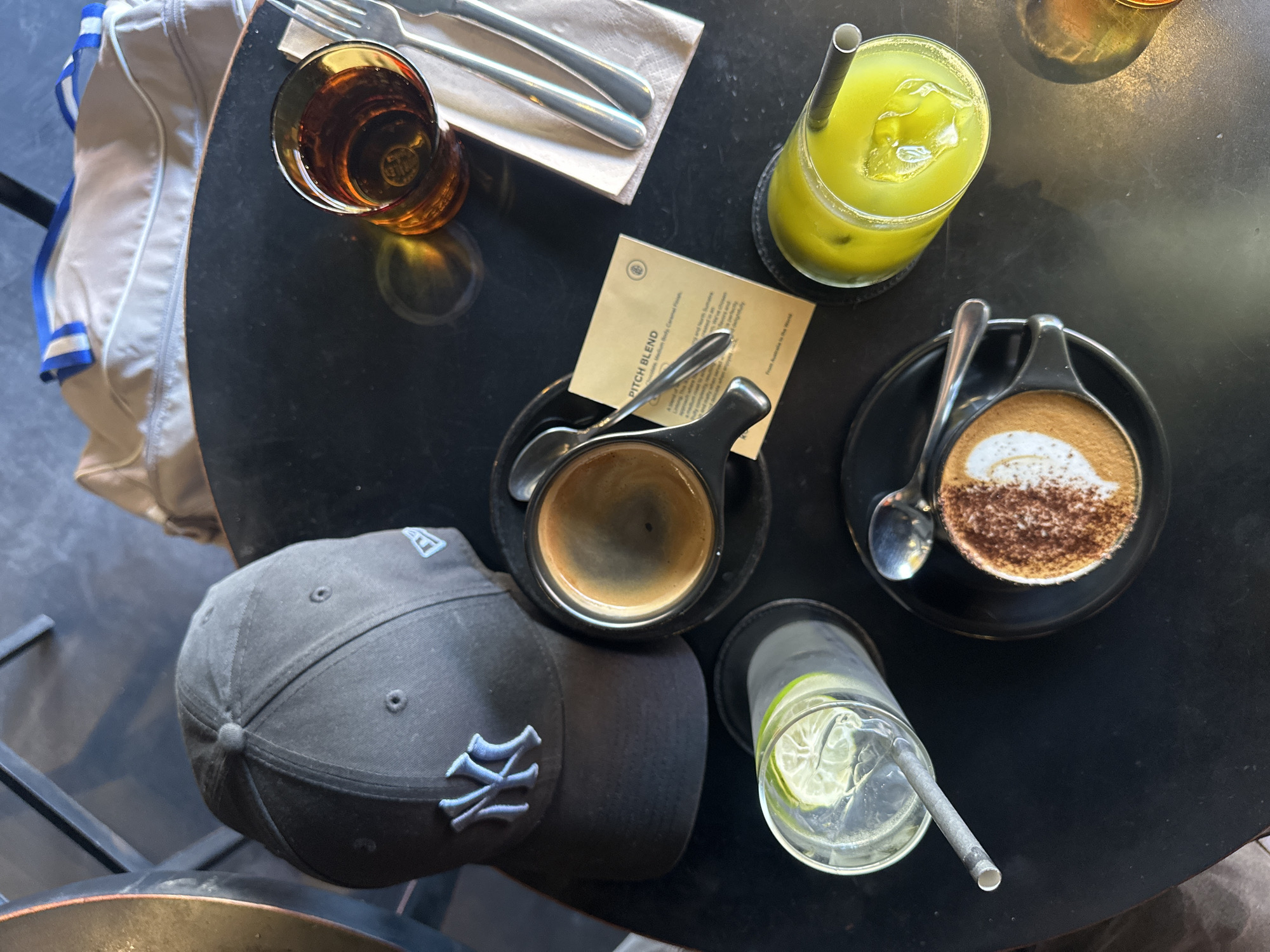 The overhead view shows a dark, round table with various items. In the upper left corner is a glass filled with a clear liquid, ice, and lime slices, with a metal straw. To its right is a black baseball cap with a blue New York Yankees logo. Below the cap is a small cup of dark coffee with crema. A small, dark saucer with a latte topped with foam and cocoa powder sits to the left of the coffee cup, with a spoon resting on the saucer. A green beverage with ice and a metal straw is in a glass to the lower left. To the right of the green drink is an amber glass with a dark liquid and a spoon. A small paper packet labeled "PITCH BLEND" is under the coffee cup, and a folded napkin with two silver utensils is to the lower right of the green drink. A tan bag with blue and white trim is visible on the right side of the image.