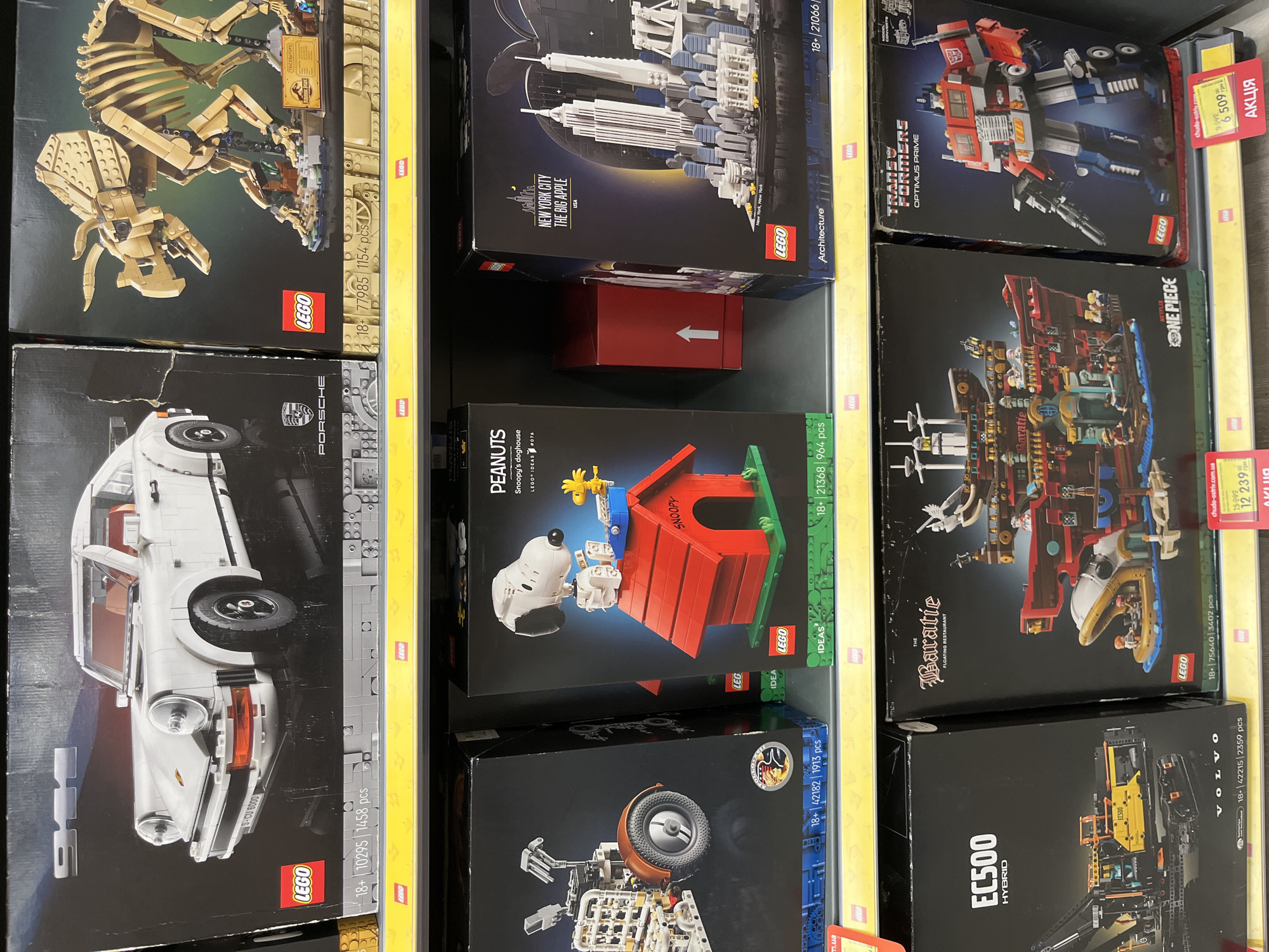 The image shows a retail shelf display of LEGO sets. On the top shelf, there are three large boxes. The leftmost box features a white Porsche 911 LEGO set. To its right is a LEGO dinosaur skeleton set, identified as a Triceratops from Jurassic Park. The rightmost box on the top shelf is not fully visible but appears to be another large LEGO set.
The middle shelf displays four more LEGO sets. From left to right, there is a LEGO Technic set with a vehicle that has a large wheel and crane. Next is a LEGO Ideas set featuring Snoopy's doghouse from Peanuts. To the right of Snoopy's doghouse is a LEGO Architecture set of New York City, showing the skyline with the Twin Towers. A red box with a white arrow is partially visible between the Snoopy and New York City sets.
The bottom shelf showcases four additional LEGO sets. The leftmost is a LEGO Technic EC500 Hybrid set. Next to it is a large LEGO set of a pirate ship called "The Baratie" from Netflix's One Piece. To the right of the pirate ship is a LEGO Transformers Optimus Prime set. A yellow price tag with red text reading "АКЦІЯ" (which means "Sale" in Ukrainian) is visible on the rightmost set and on the set to its left.
The background appears to be a store environment with other shelves and displays, suggesting a toy store or a department store section dedicated to toys. The overall lighting is bright, typical of retail displays. The image is a photograph taken at eye level, looking across the shelves. The exact location in Kharkiv, Ukraine cannot be determined from the image alone, but the presence of Ukrainian currency (implied by "UAH" on price tags) and the word "АКЦІЯ" strongly suggests a Ukrainian retail setting.