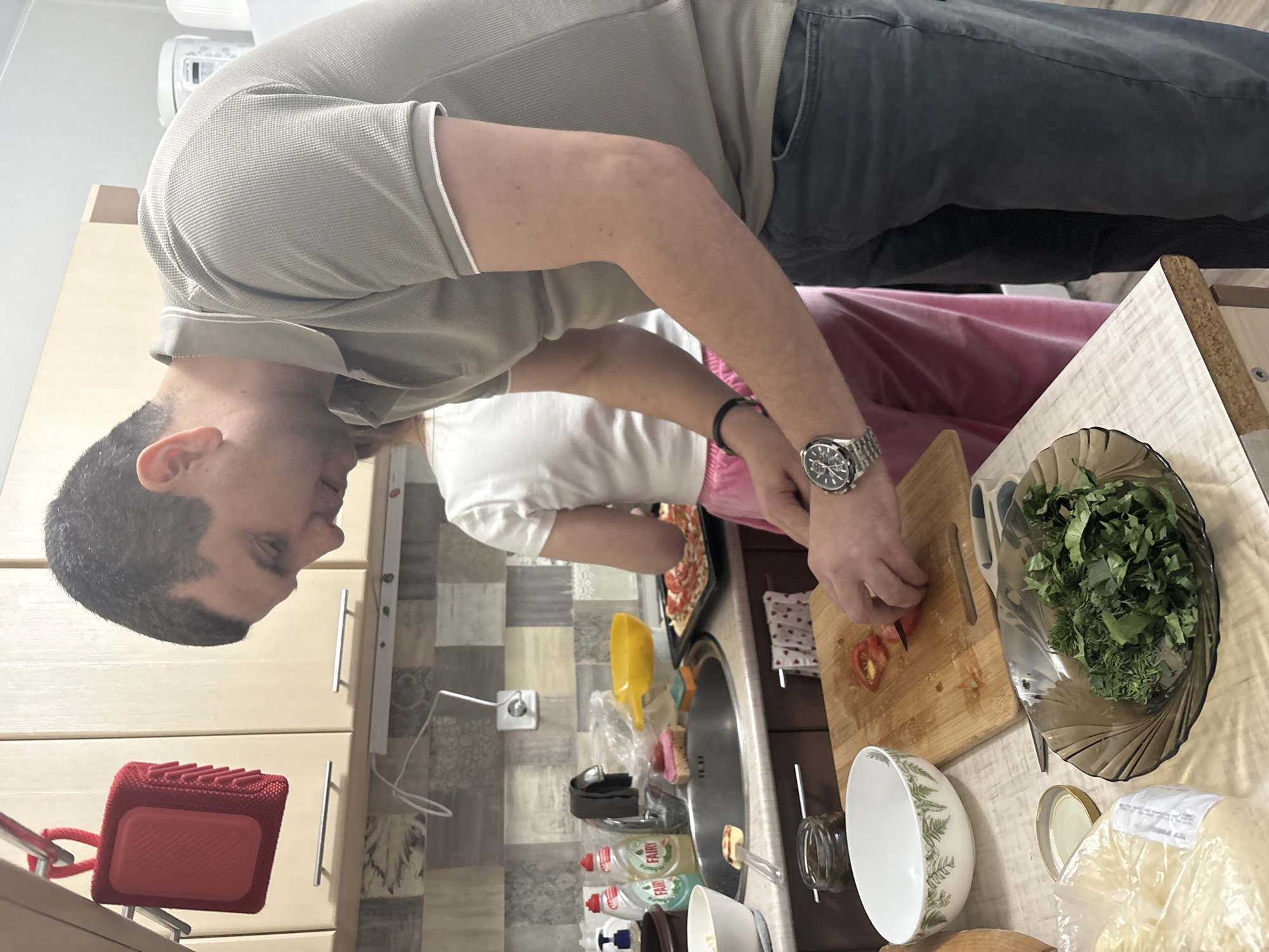 A man is cutting tomatoes on a wooden cutting board. A woman is near him and a tray with sliced tomatoes can be seen. There are bowls, a sink, and other kitchen items on the counter. The location is Кондратово, Russia.