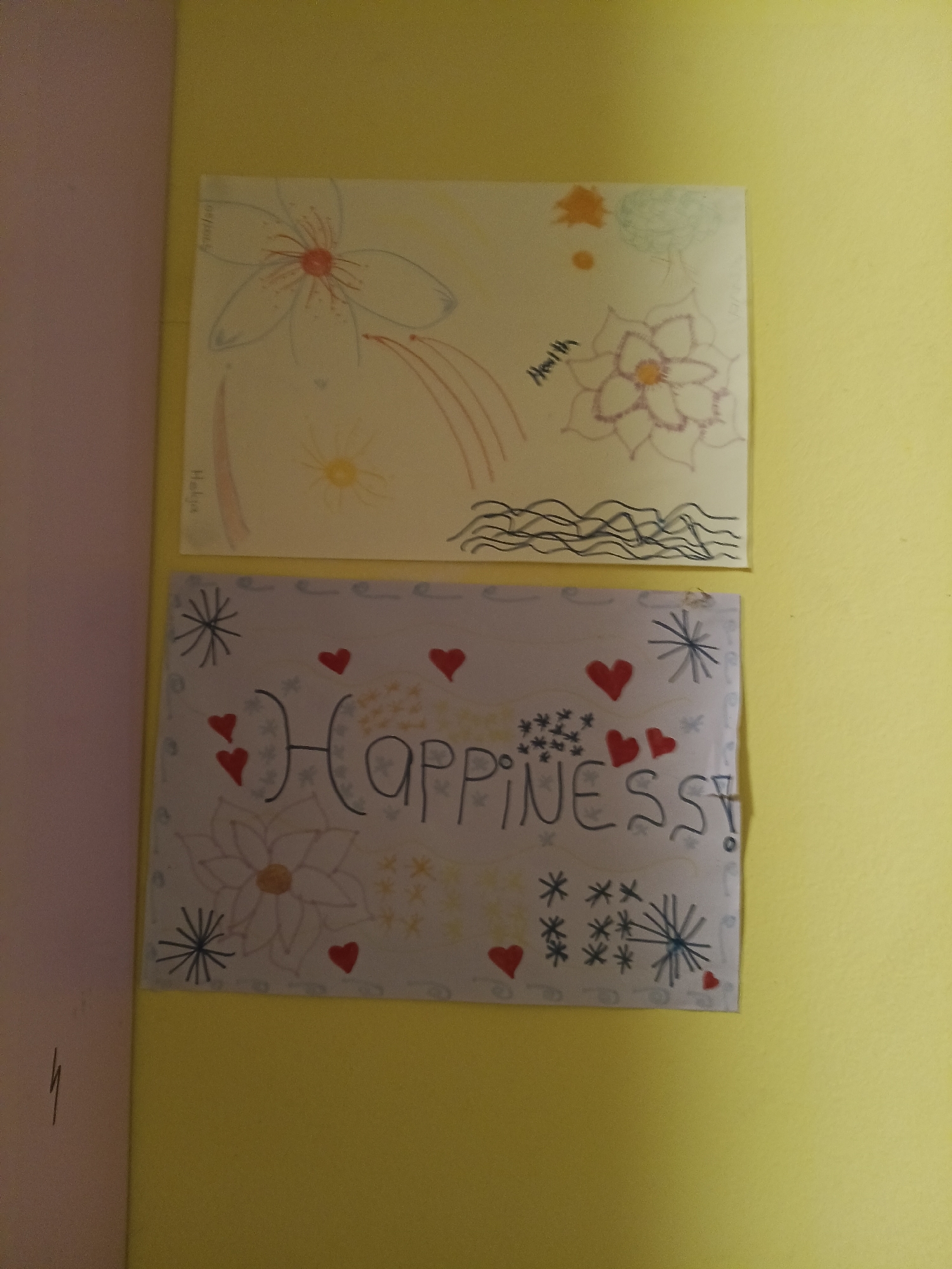 The image shows two drawings on white paper attached to a yellow wall. The top drawing features several hand-drawn flowers, some with red and orange centers, and what appears to be a rainbow or streaks of color. There is also a section of wavy lines at the bottom of this drawing. The word "Health" is written in cursive on this drawing. The bottom drawing contains the word "HAPPINESS!" written in large, dark letters. This drawing also includes several red hearts, small snowflake-like shapes, and a larger flower design in the center. The drawings are attached to the wall with pins or staples. The overall scene is domestic, likely a child's artwork displayed on a wall. There is no information in the image that can place it in Wels, Austria.