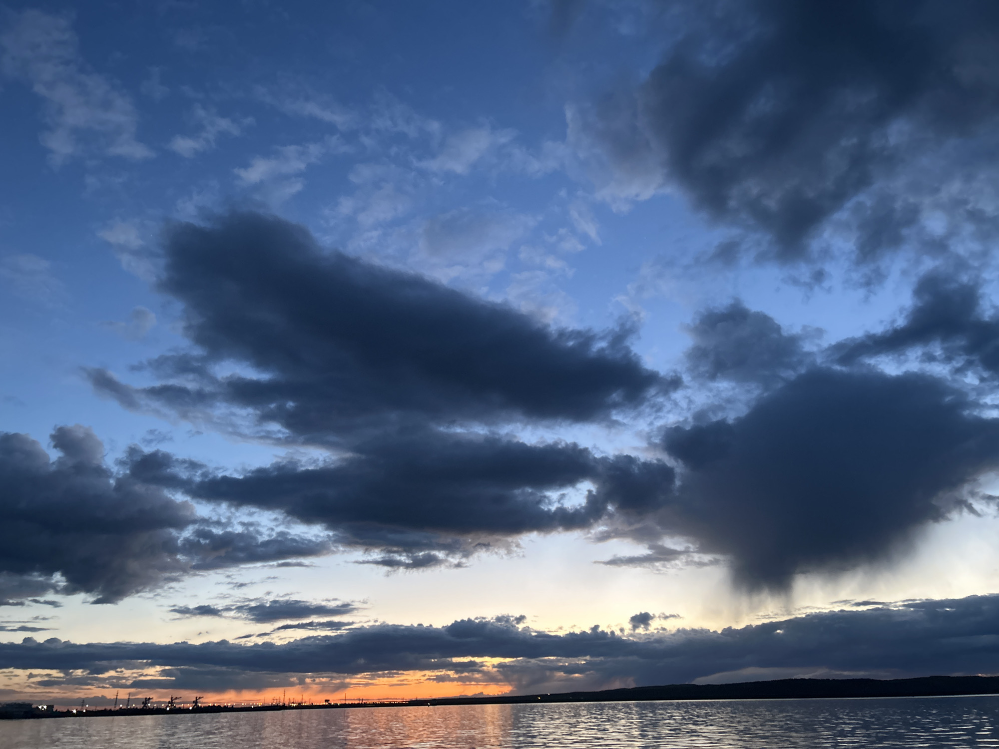 The image shows a body of water, likely a lake or a wide river, with a shoreline visible in the distance. On the shoreline, there are silhouettes of structures, possibly industrial or port facilities with cranes, suggesting a cityscape. The sky is filled with a mix of dark, heavy clouds and patches of blue. In one area, rain or precipitation is visible falling from a cloud. The overall lighting indicates it's either sunset or sunrise. The location is plausibly Chaikovsky, Russia, given its industrial features on the shore and the broad river or lake setting.