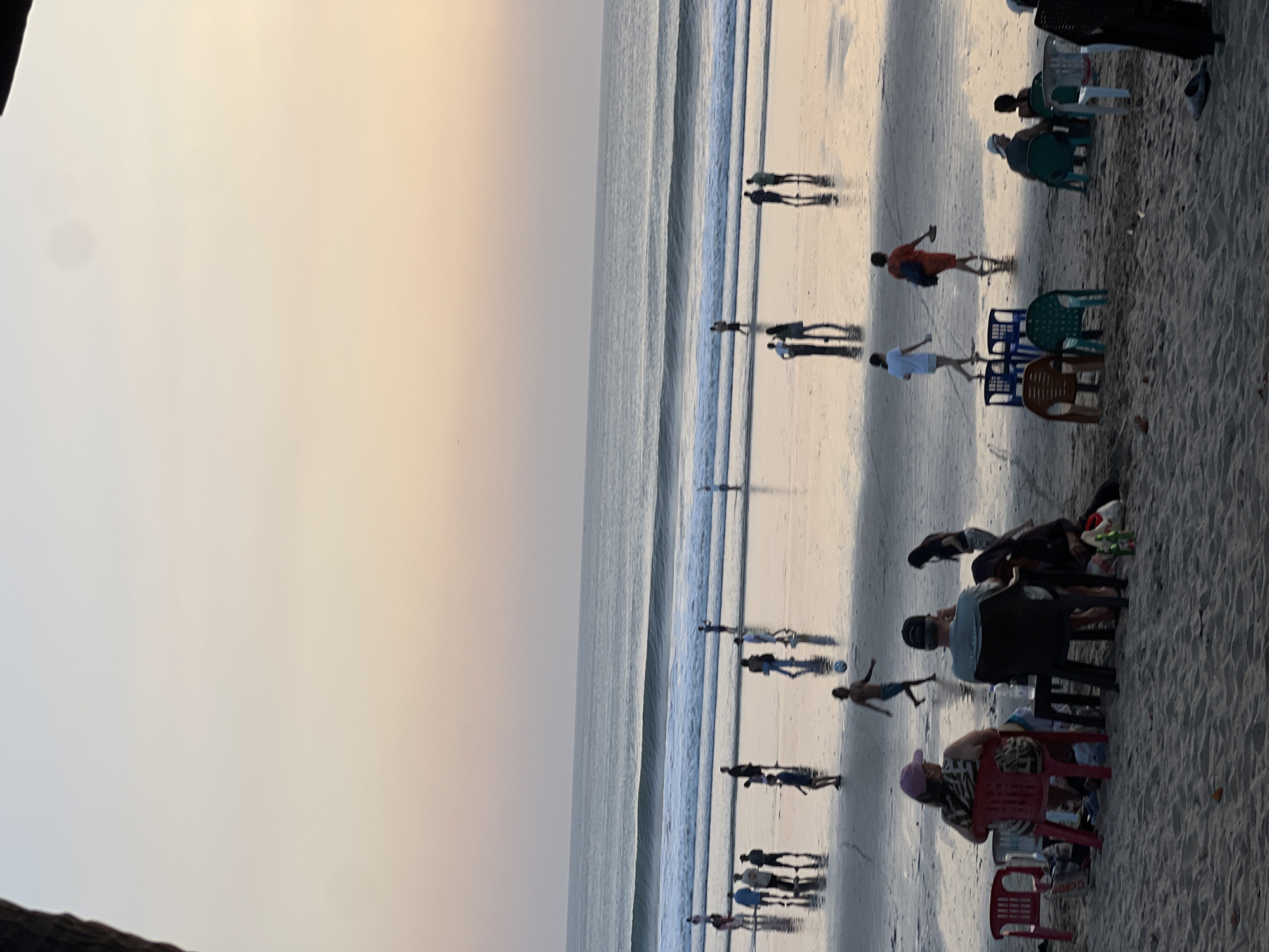 This is a photograph taken at dusk on Kuta Beach, Bali, Indonesia. The scene captures a lively beach environment with numerous people engaged in various activities. In the foreground, a few individuals are seated on chairs on the sandy shore, some appearing to be having a meal or drinks, with tables and bags nearby. Further out on the wet sand, more people are walking and interacting, their reflections visible in the shallow water. A group of individuals are playing soccer, with one person in motion to kick a ball. The ocean in the background features gentle waves breaking on the shore, creating a foamy white line. The sky above is a gradient of soft pastel colors, transitioning from a pale orange near the horizon to a light blue, indicating the setting of the sun. The overall atmosphere is one of leisure and recreation.