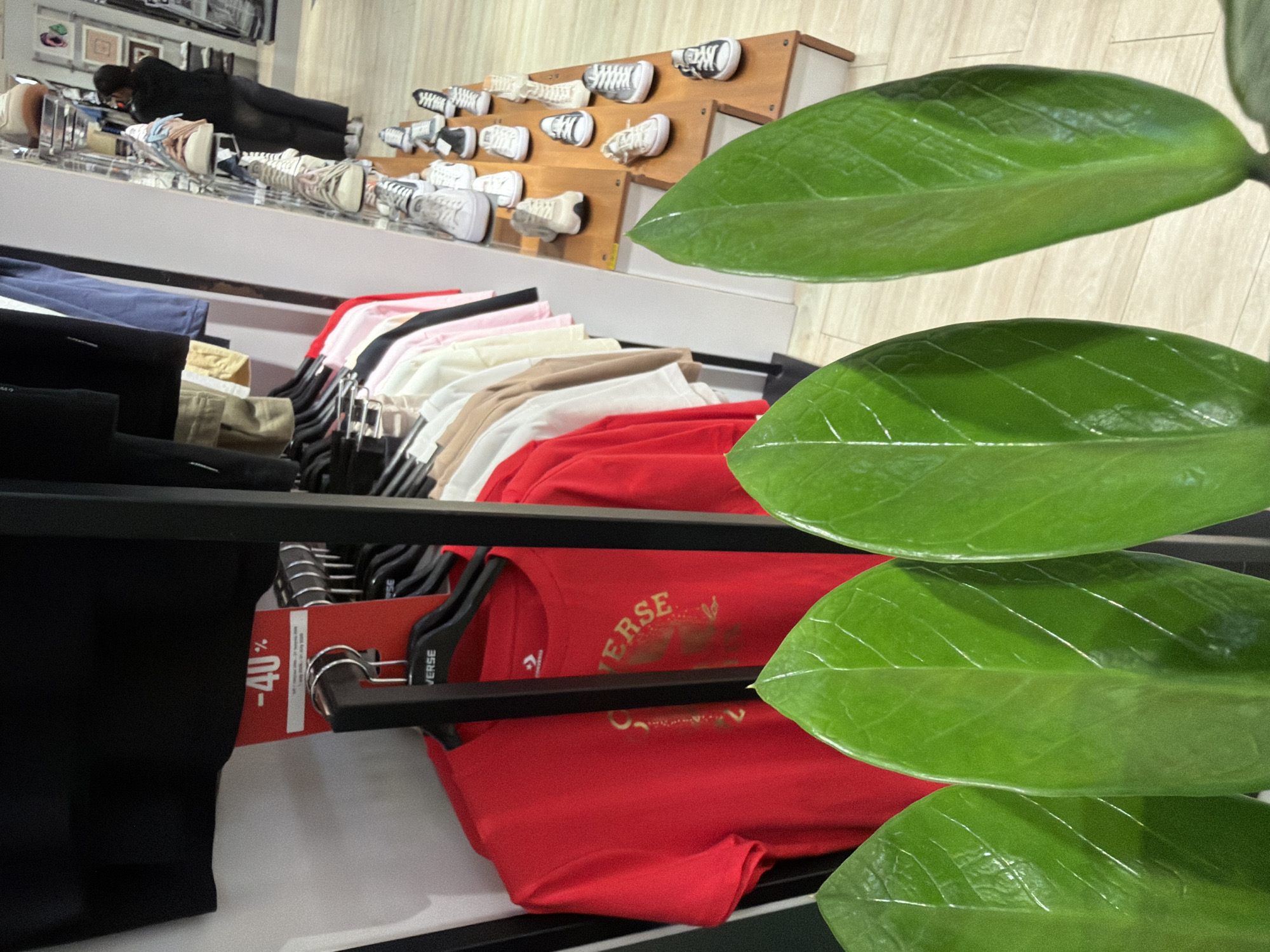 An indoor clothing store, likely a Converse outlet, is depicted. In the foreground, three large, glossy green leaves obscure the bottom portion of the image. Behind the leaves, a rack of Converse t-shirts is visible, with a bright red t-shirt prominently displayed on a hanger with the Converse logo. A " -40% " sale tag is attached to the rack. Further in the background, a tiered display of Converse sneakers lines a shelf. A woman, dressed in dark clothing and a face mask, is seen browsing the sneaker display. The store appears well-lit with light-colored flooring and shelving. The location is identified as Ko Samui District, Thailand.