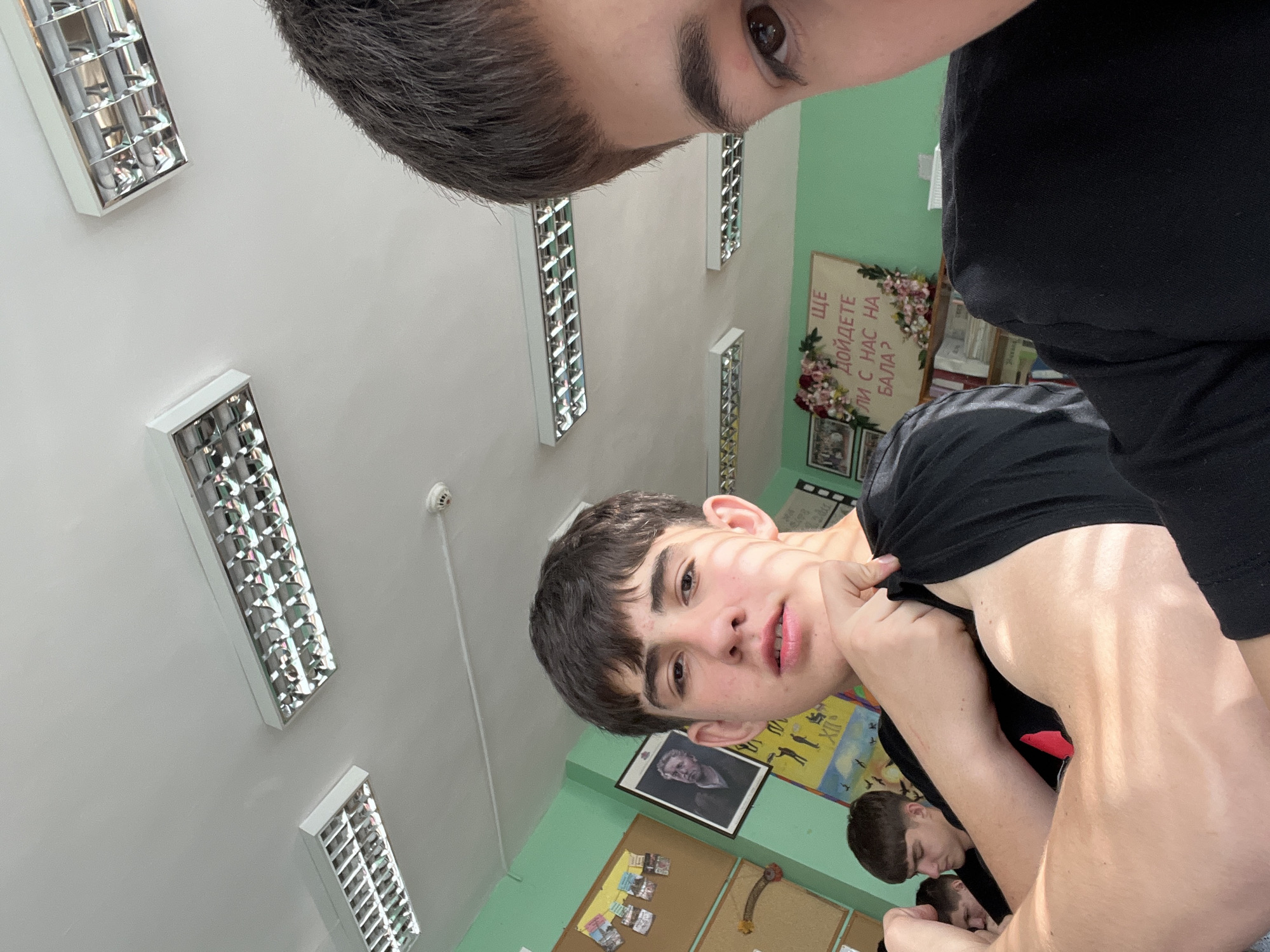 The image shows three young men in an interior setting, possibly a classroom or office, due to the fluorescent lights, bulletin board, and poster visible in the background. The setting appears to be indoors. The faces of two men are prominently visible, one closer to the camera and partially out of frame. The man closer to the camera seems to be holding the arm of the other. Another man is partially visible in the lower center of the frame. The location might be in Asenovgrad, Bulgaria.